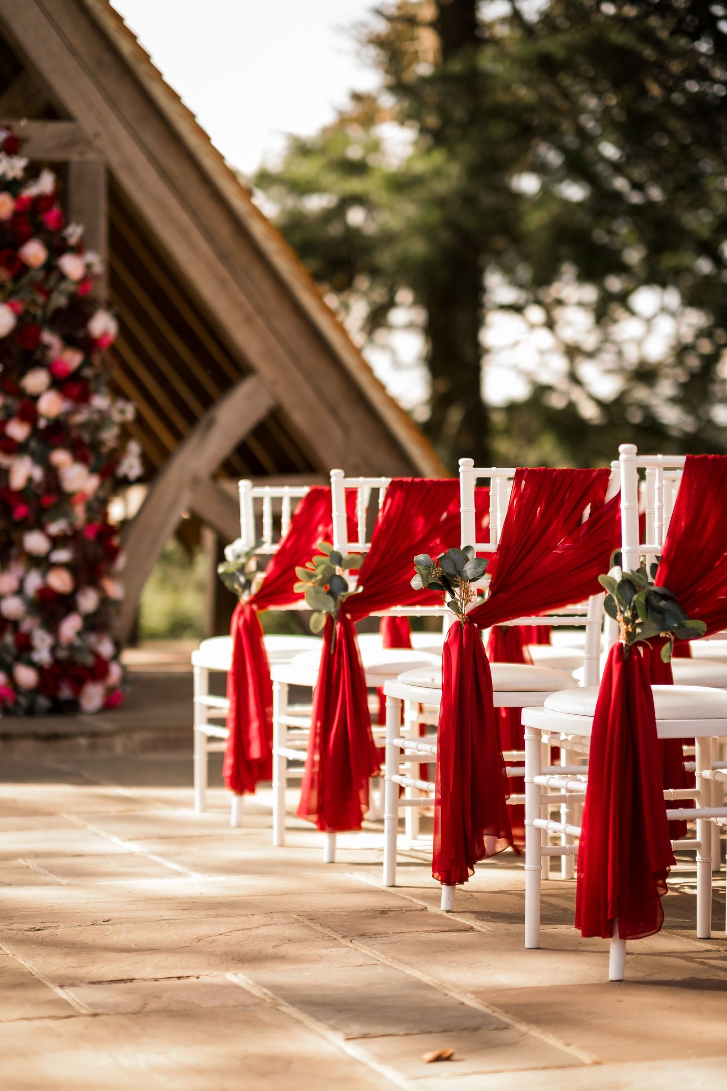 White chairs adorned with red bows and floral accents line up for an elegant outdoor wedding ceremony setup.