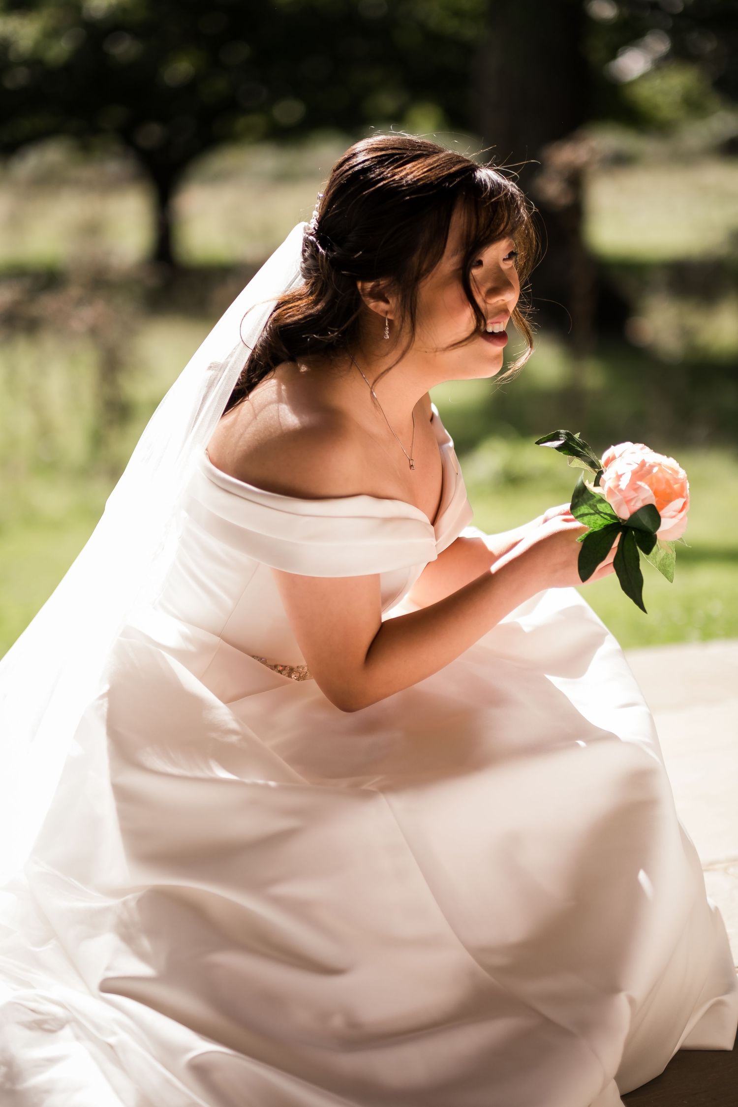 A bride in an off-shoulder white wedding gown sits outdoors holding a peach rose in soft natural lighting.