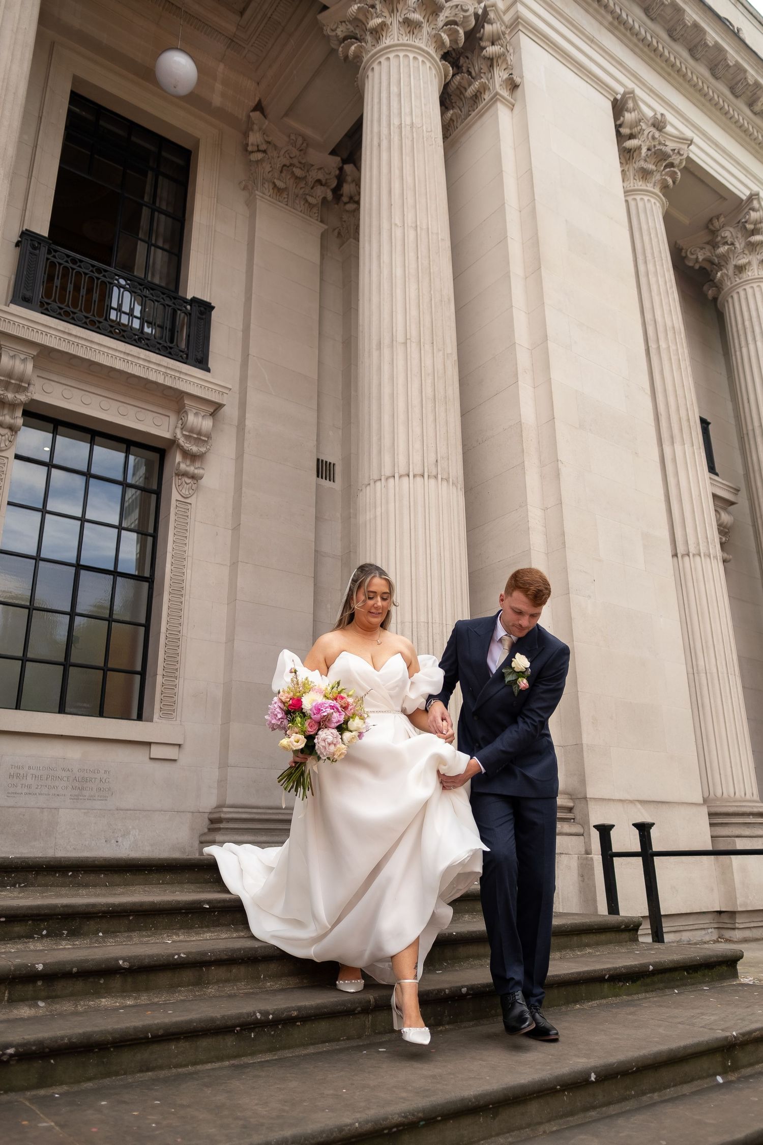 Bride and groom descend grand stone steps with columns, she holds a pink bouquet, he wears a navy suit.