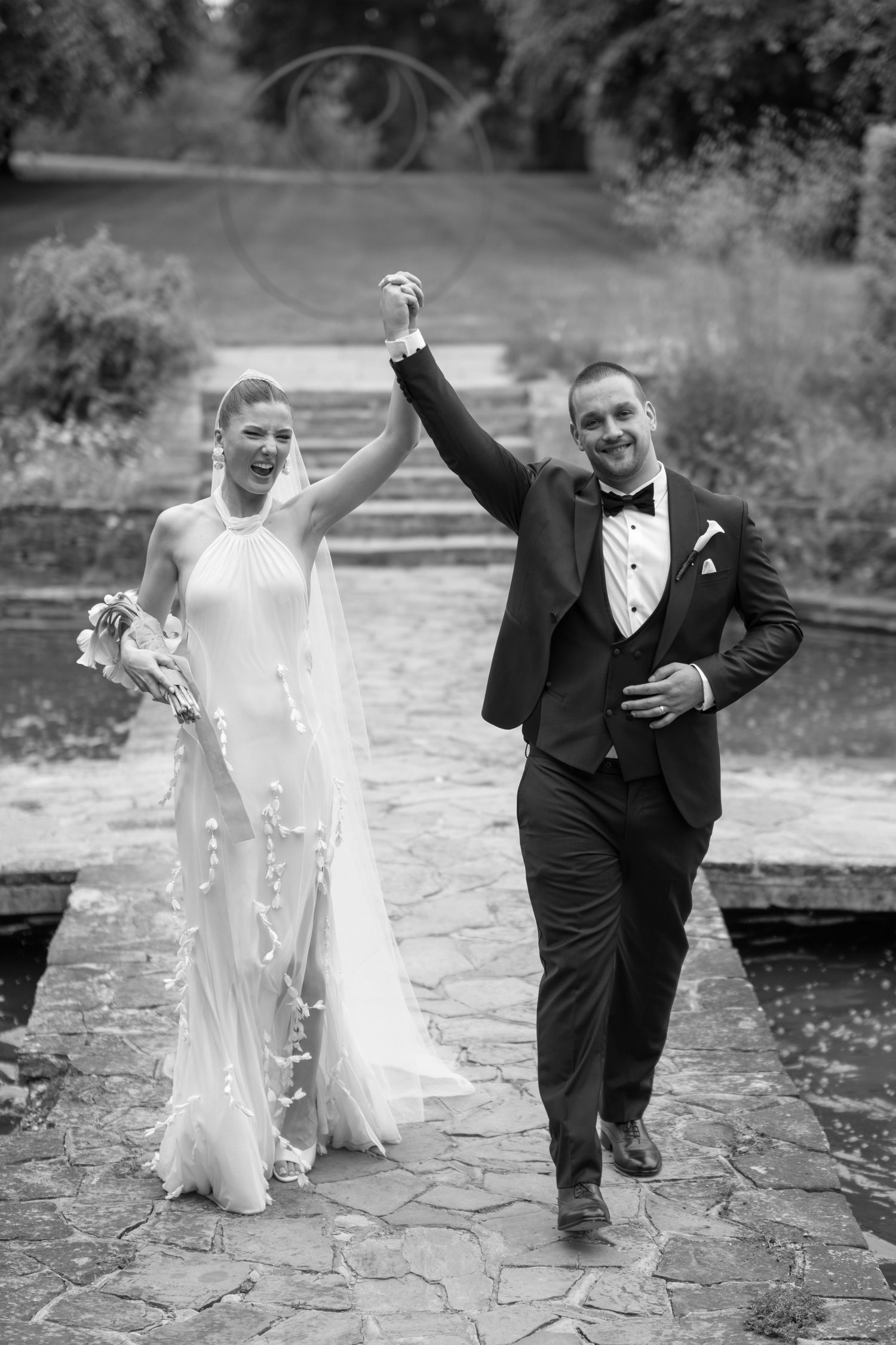 Black and white photo of joyful newlyweds celebrating, bride in elegant gown, groom in tuxedo, raising hands triumphantly outdoors.