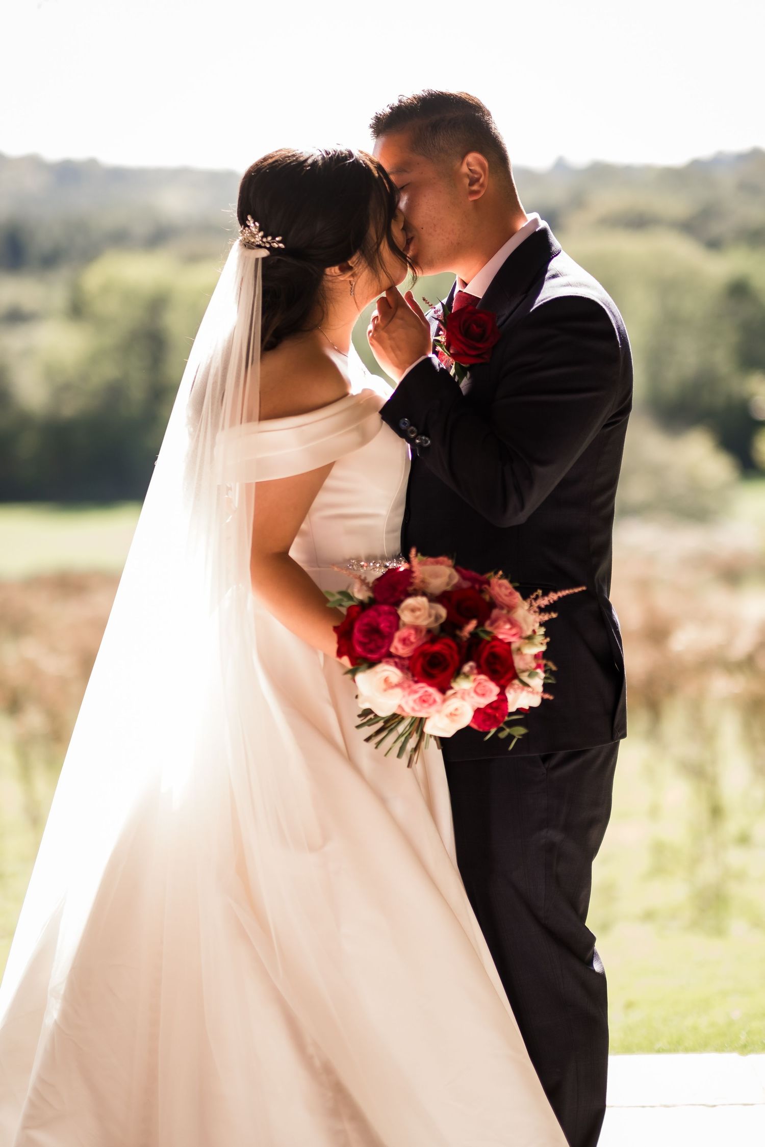 A romantic wedding couple shares a tender moment outdoors with a beautiful red bouquet and flowing white veil.