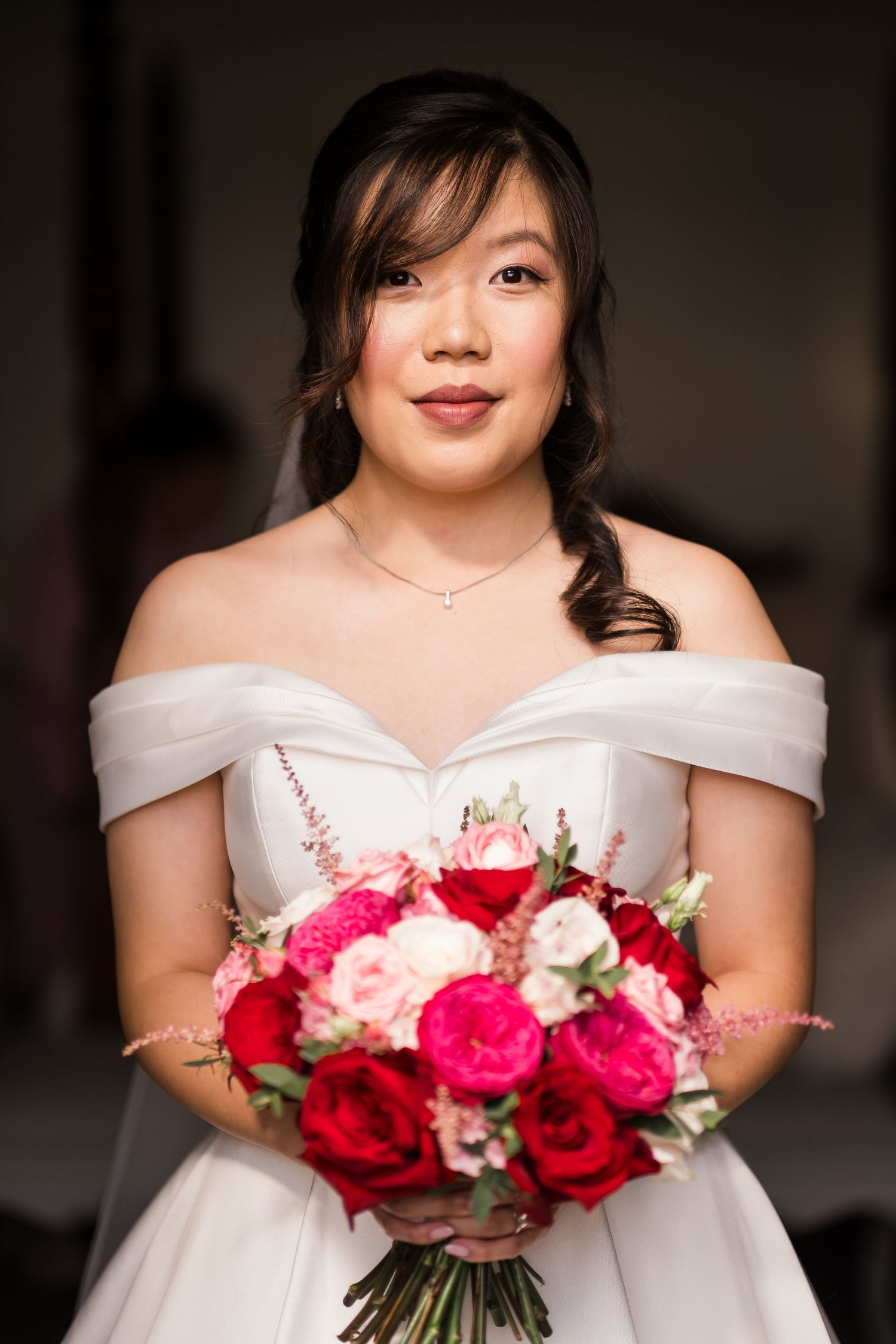 Elegant bride in off-shoulder white gown holds vibrant red and pink rose bouquet against dark background.