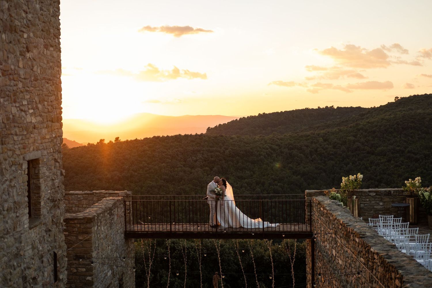 Bride and groom share a romantic kiss on a stone balcony at sunset overlooking lush green hills in Tuscany.