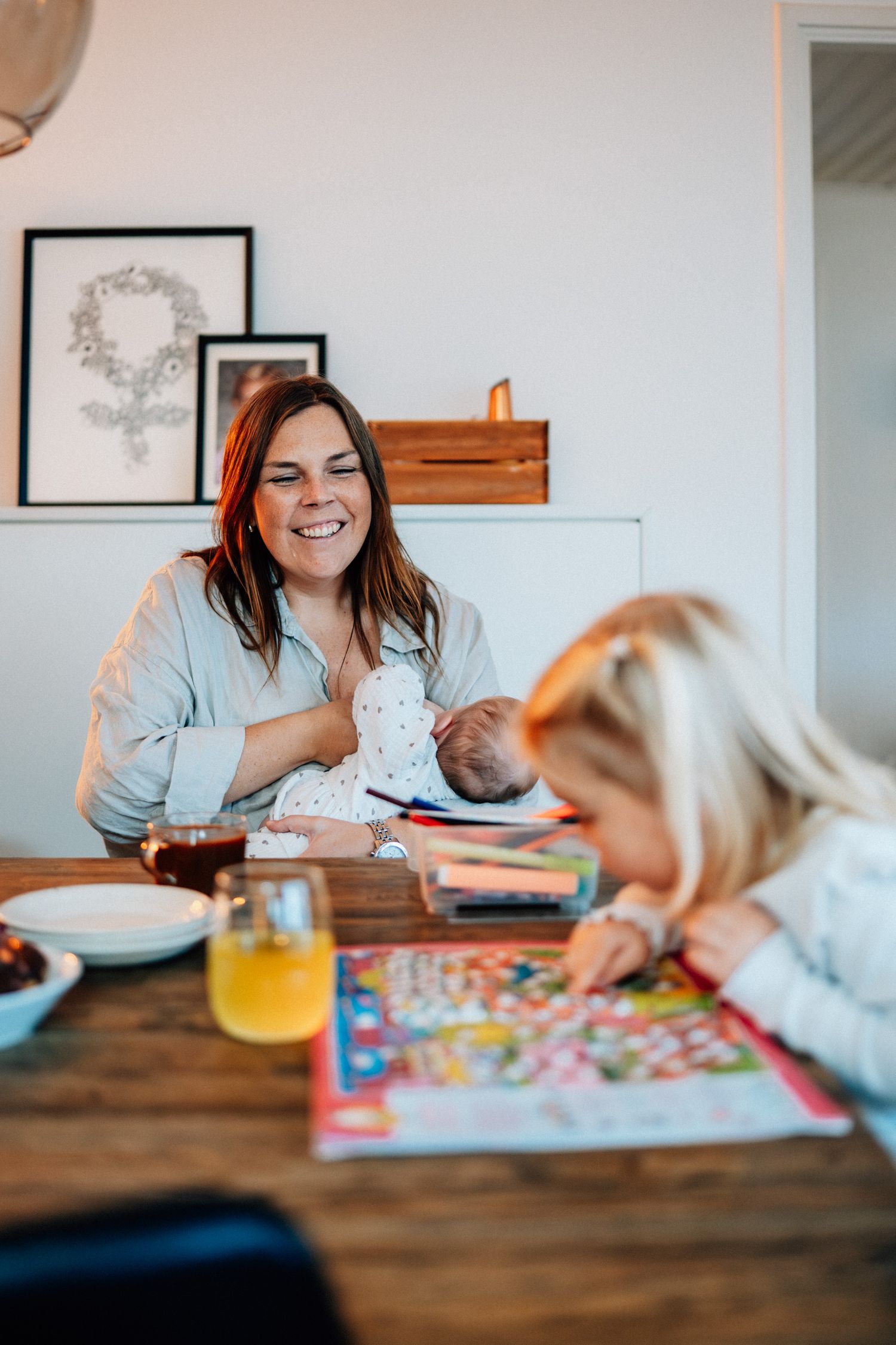 Vid ett bort sitter en mamma med en bebis i knät och ler åt storasyster som pysslar.