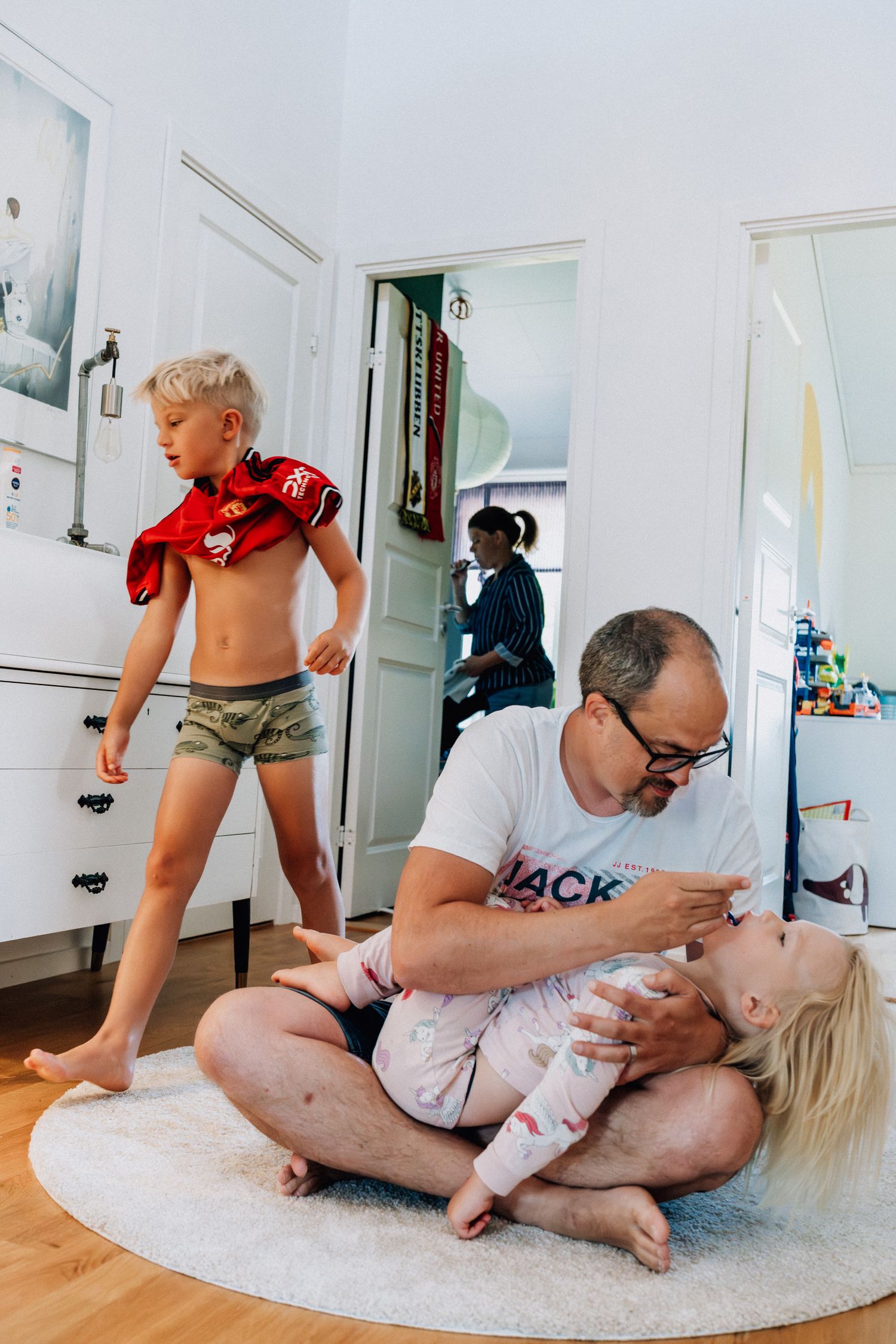 En pappa borstar tänderna på sitt barn samtidigt som ett annat bar går förbi halvklätt och en mamma står i bakgrunden och borstar tänderna.