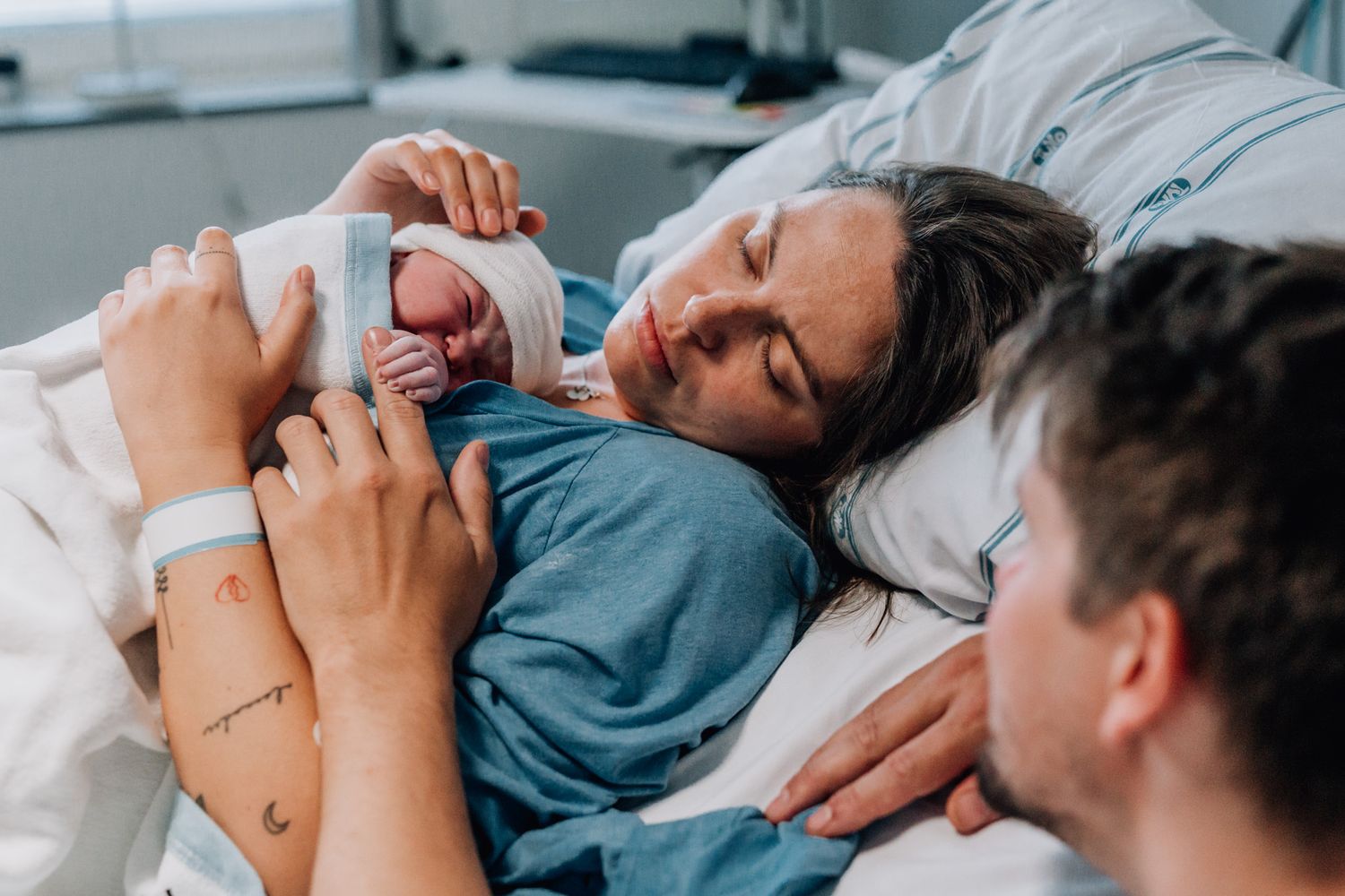 Mamman ligger i förlossningssängen med sin nyfödda bebis på magen och pappan sitter bredvid.