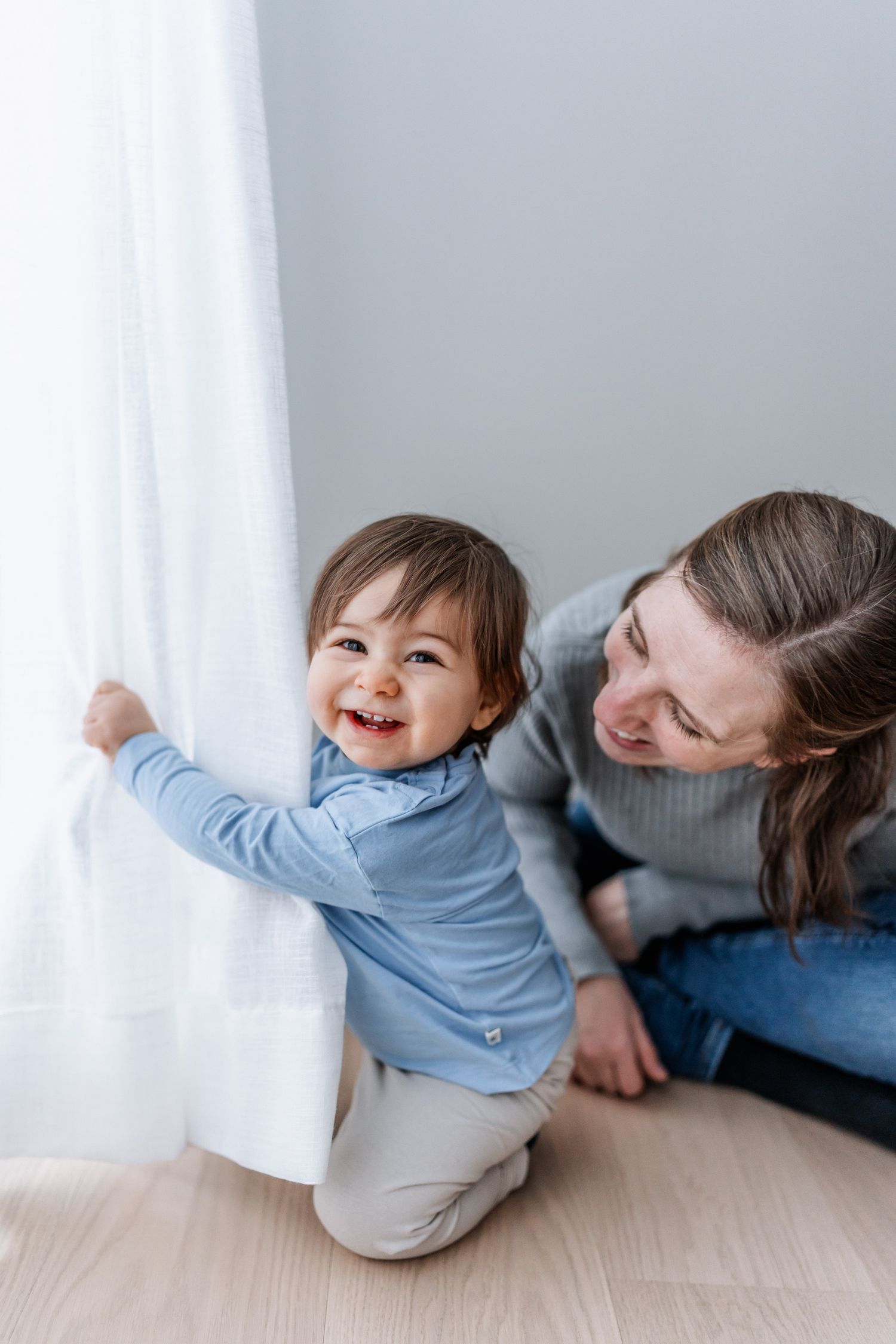 Det lilla barnet tittar busigt fram bakom gardinen och har sin mamma bredvid sig.