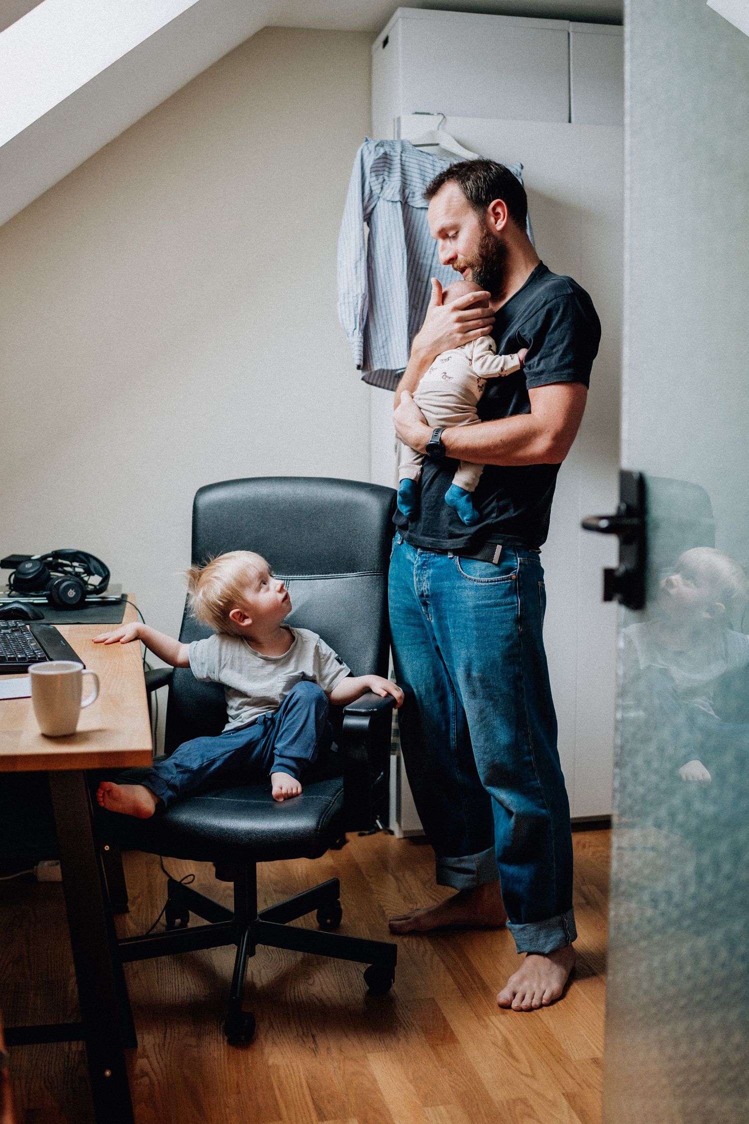 Sonen sitter på en kontorsstol och tittar upp på pappan som håller i en bebis.