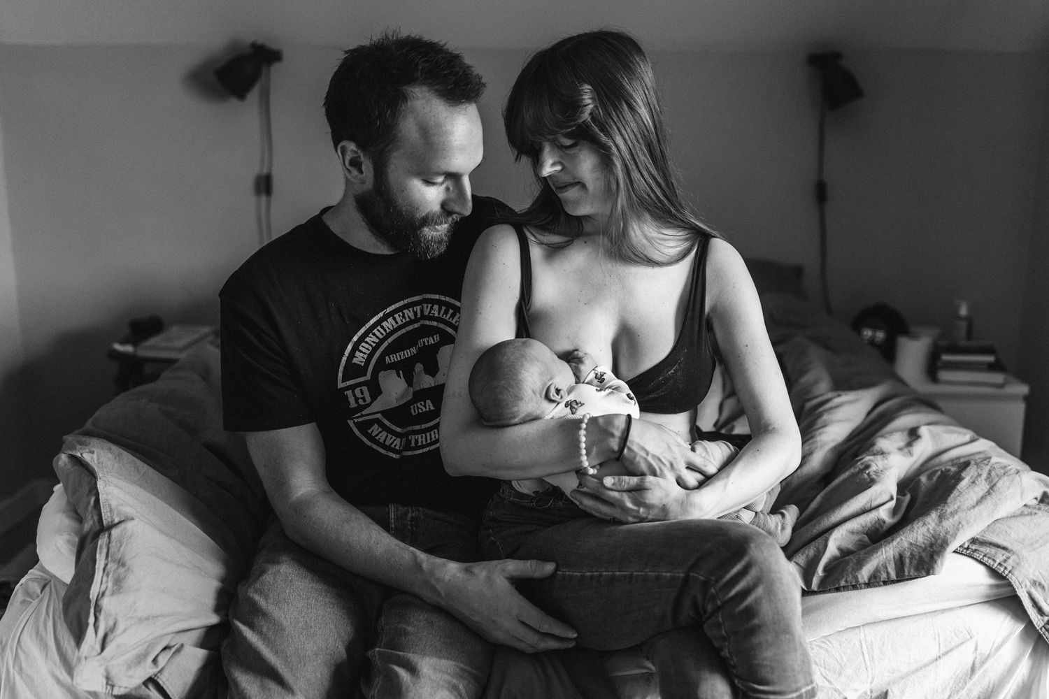 En mamma sitter på en sängkant och ammar sin bebis och pappan håller om dem.