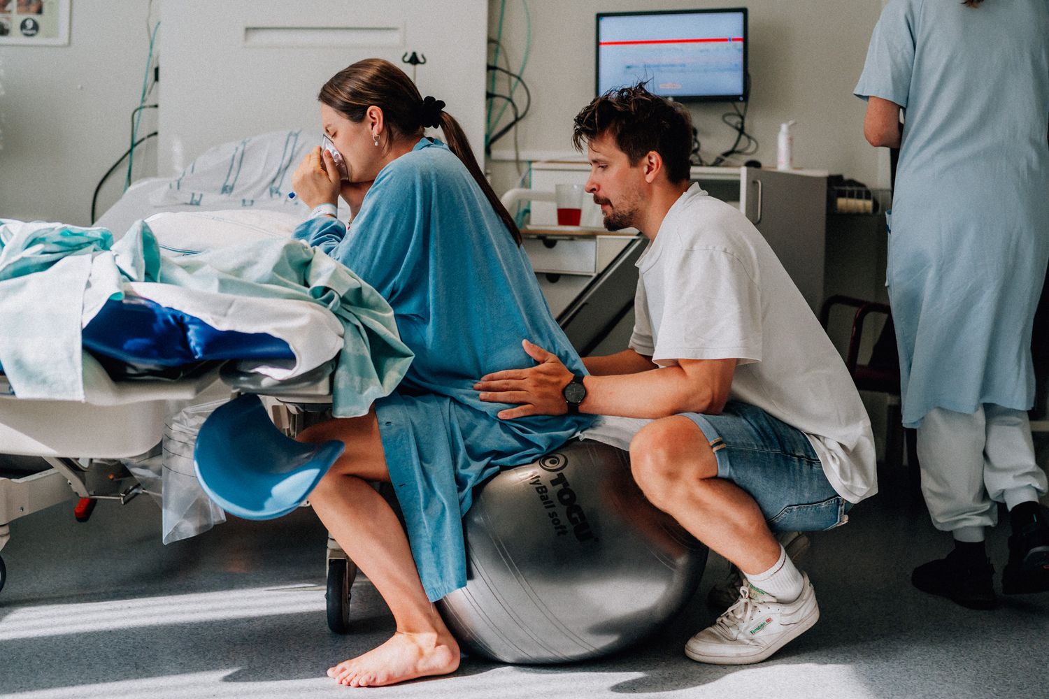 En gravid kvinna sitter på en pilatesboll i förlossningsrummet med en andningsmask och hennes man sitter bakom och trycker på hennes höft.