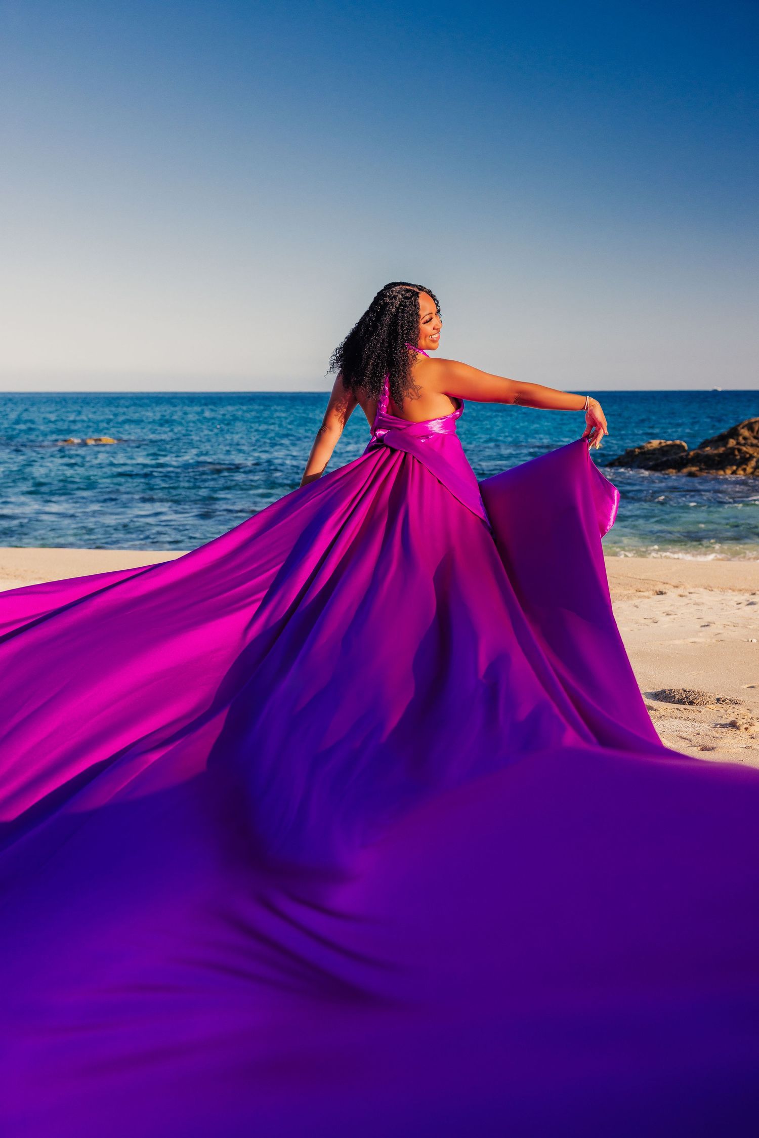 Woman in flowing purple-pink gown poses on sandy beach with ocean backdrop, fabric billowing dramatically in the breeze.
