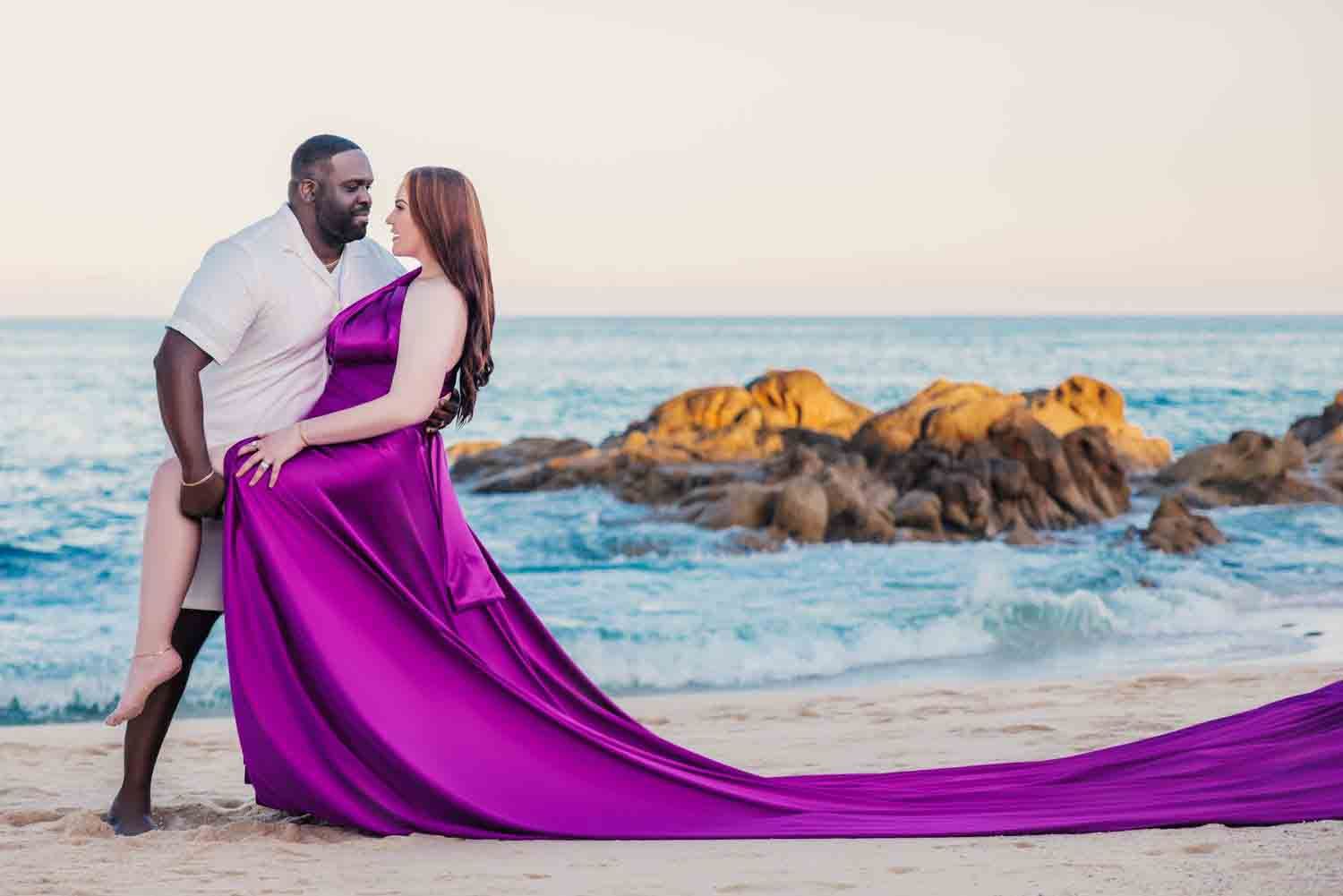 Couple embracing on a sandy beach, woman in flowing purple gown, rocky ocean backdrop at sunset.