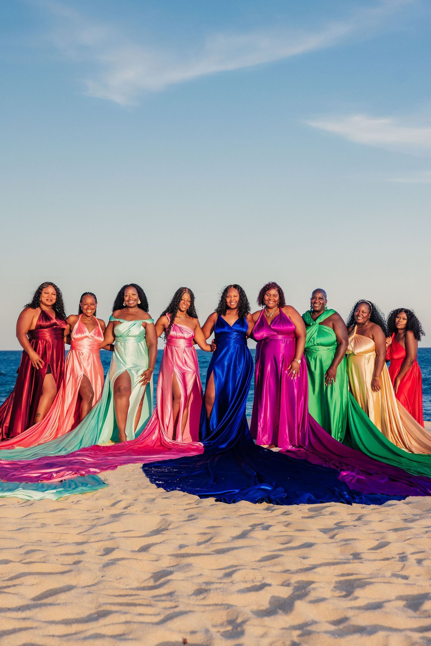 Ten women in vibrant rainbow satin gowns pose together on a sandy beach with ocean backdrop.