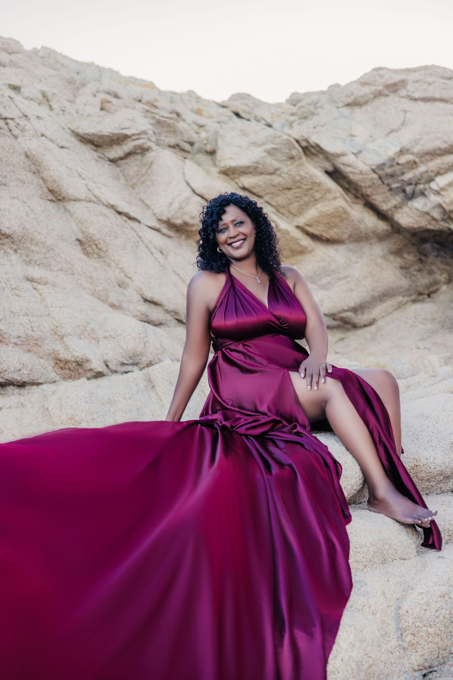 Woman in flowing burgundy satin gown sitting on rocks, smiling joyfully in an outdoor rocky landscape.