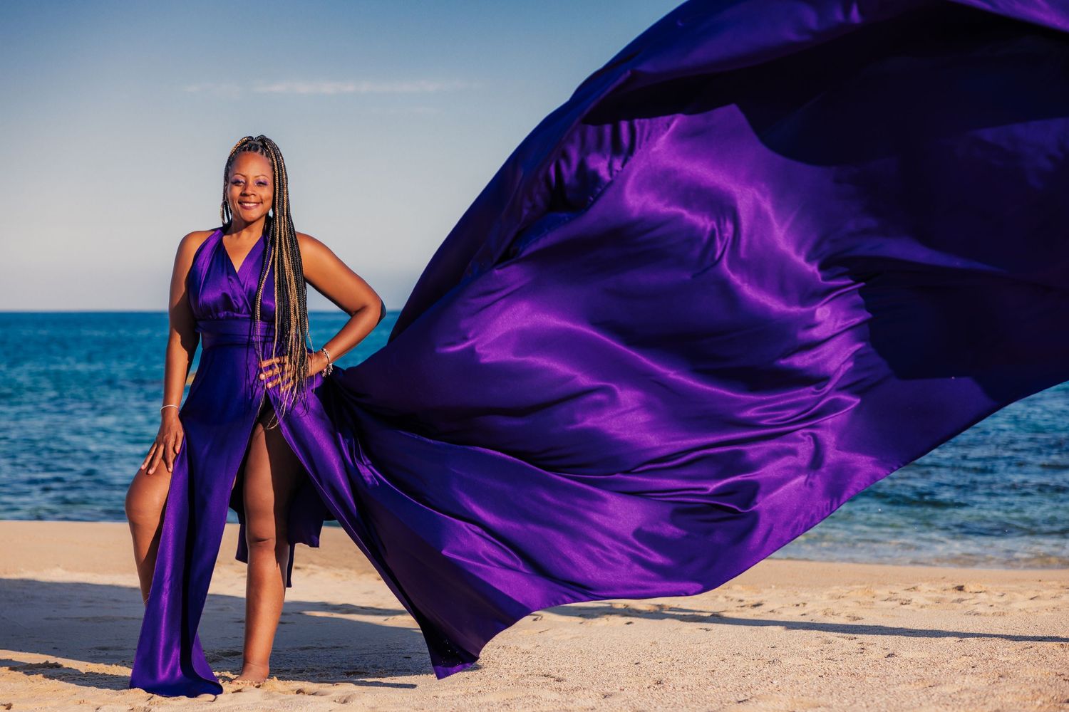 Woman in flowing purple satin gown on sandy beach, fabric billowing in wind against blue ocean sky.
