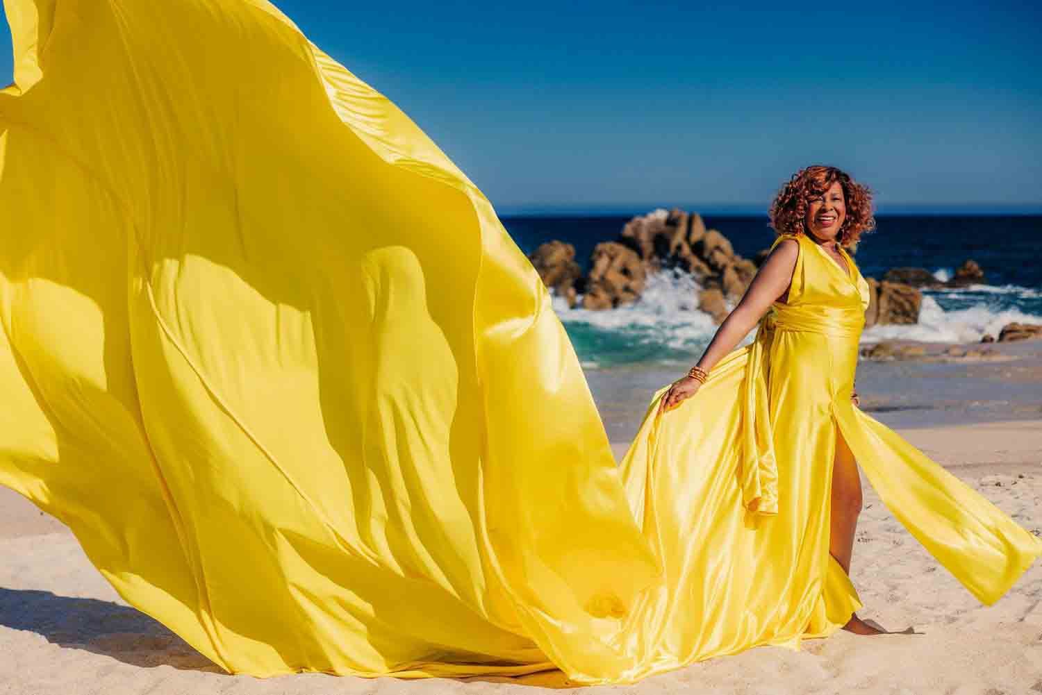 Woman in flowing yellow gown on sandy beach with ocean waves and rocks in background.