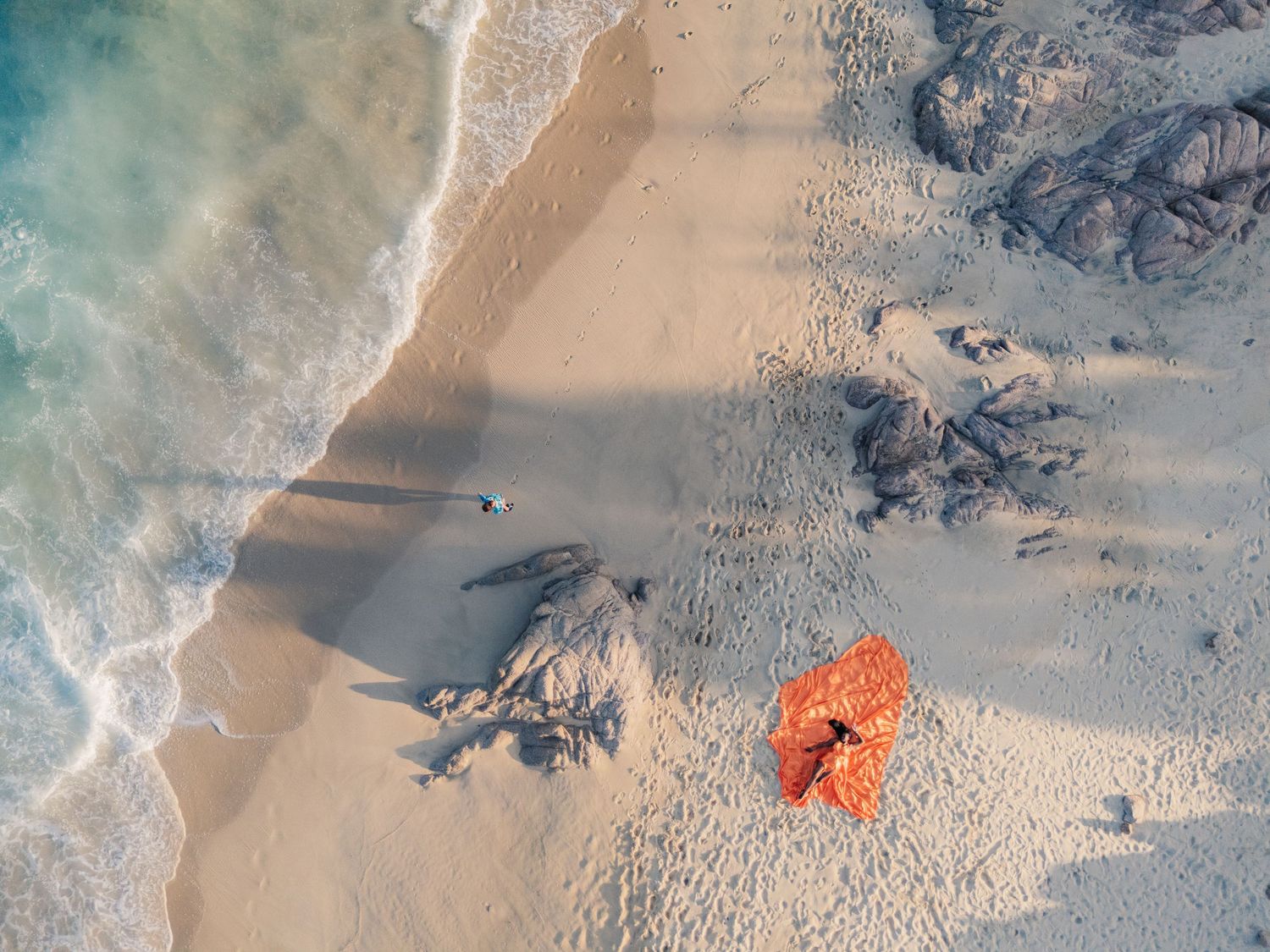 Aerial view of a white sandy beach with turquoise waves, rocks, and a person lying on an orange towel.