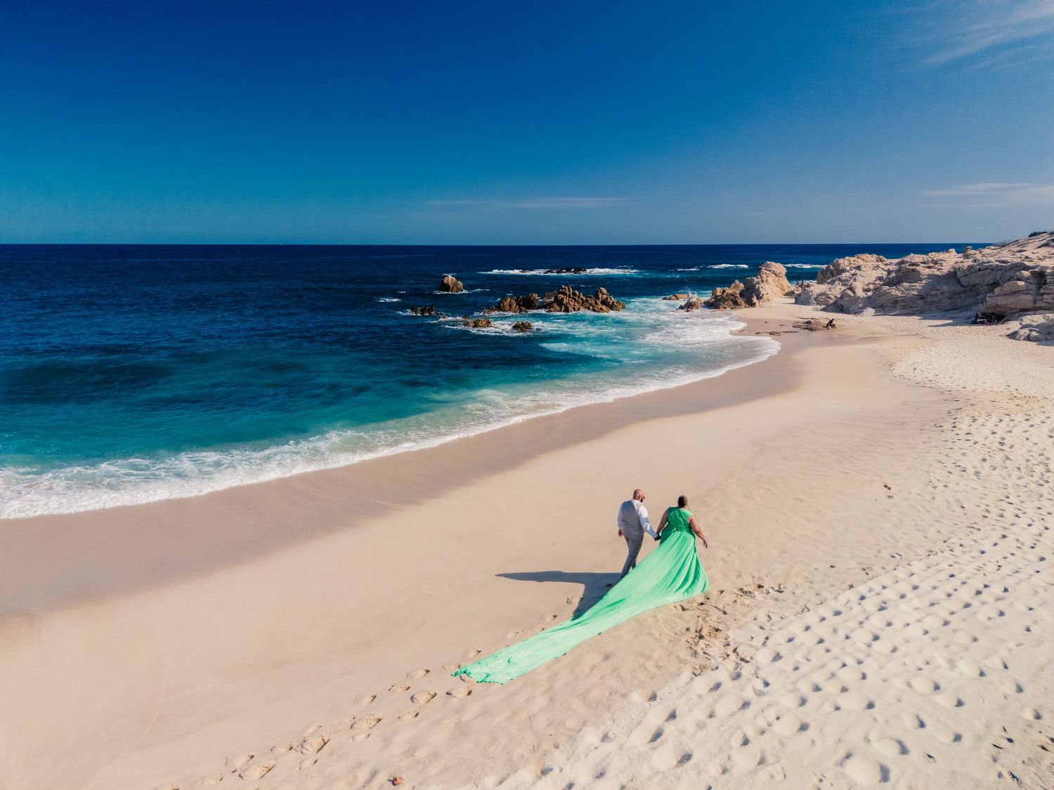Couple in green flowing dress on white sand beach with turquoise ocean and rocky outcrops under clear blue sky.