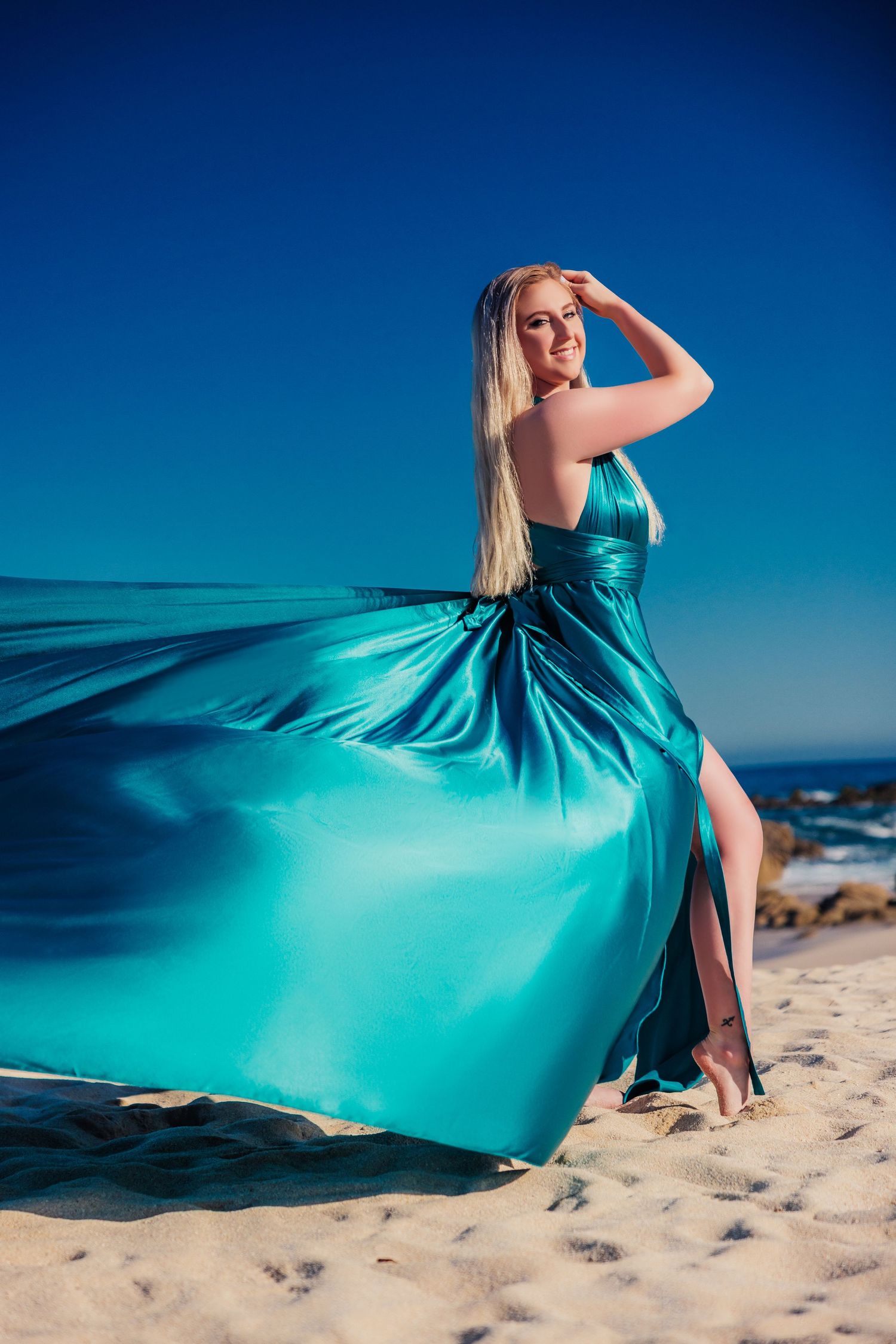 Blonde woman in flowing teal satin gown on sandy beach, dress billowing in wind against deep blue sky.