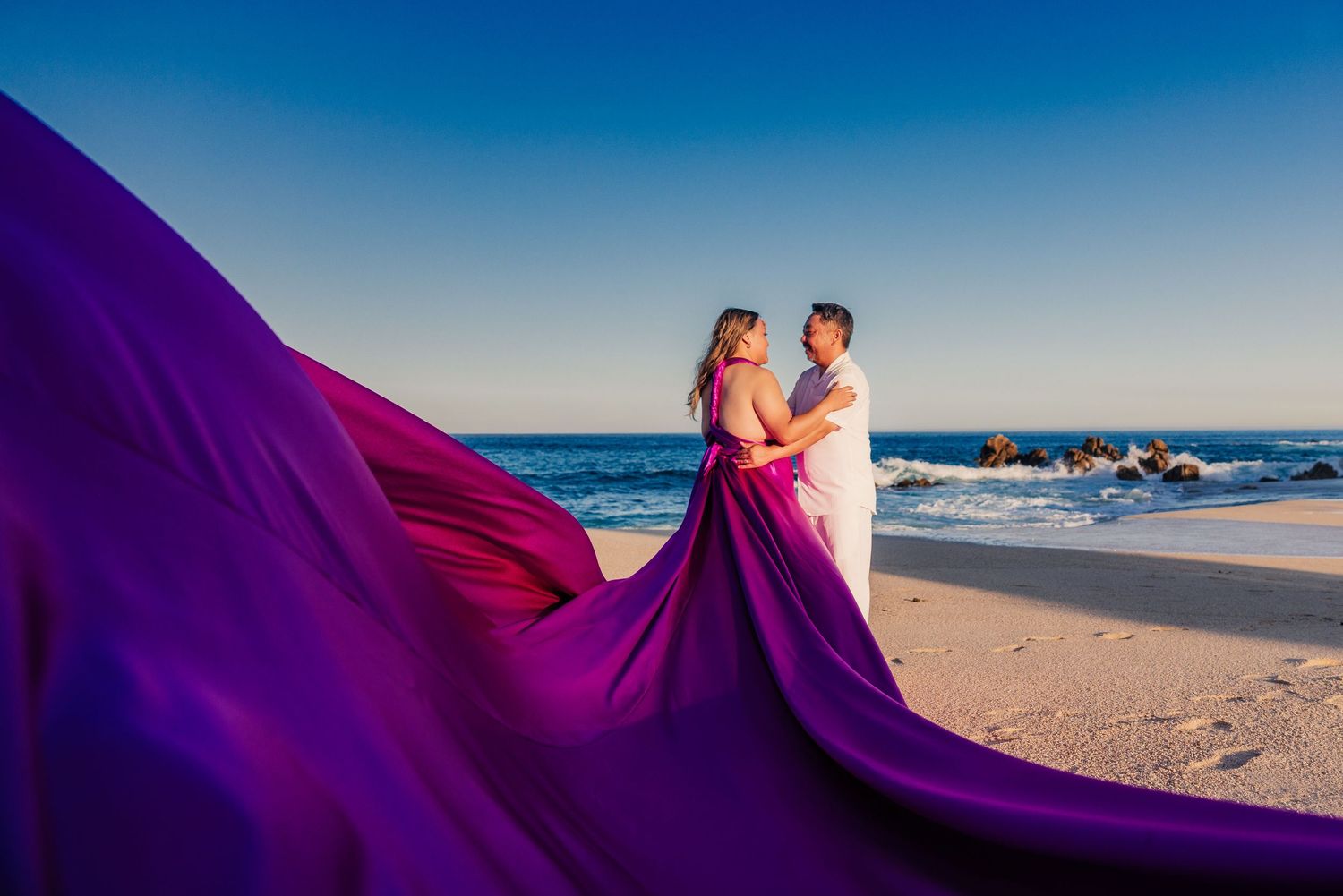 Couple embracing on a beach with a flowing purple gown billowing in the wind against a clear blue sky.
