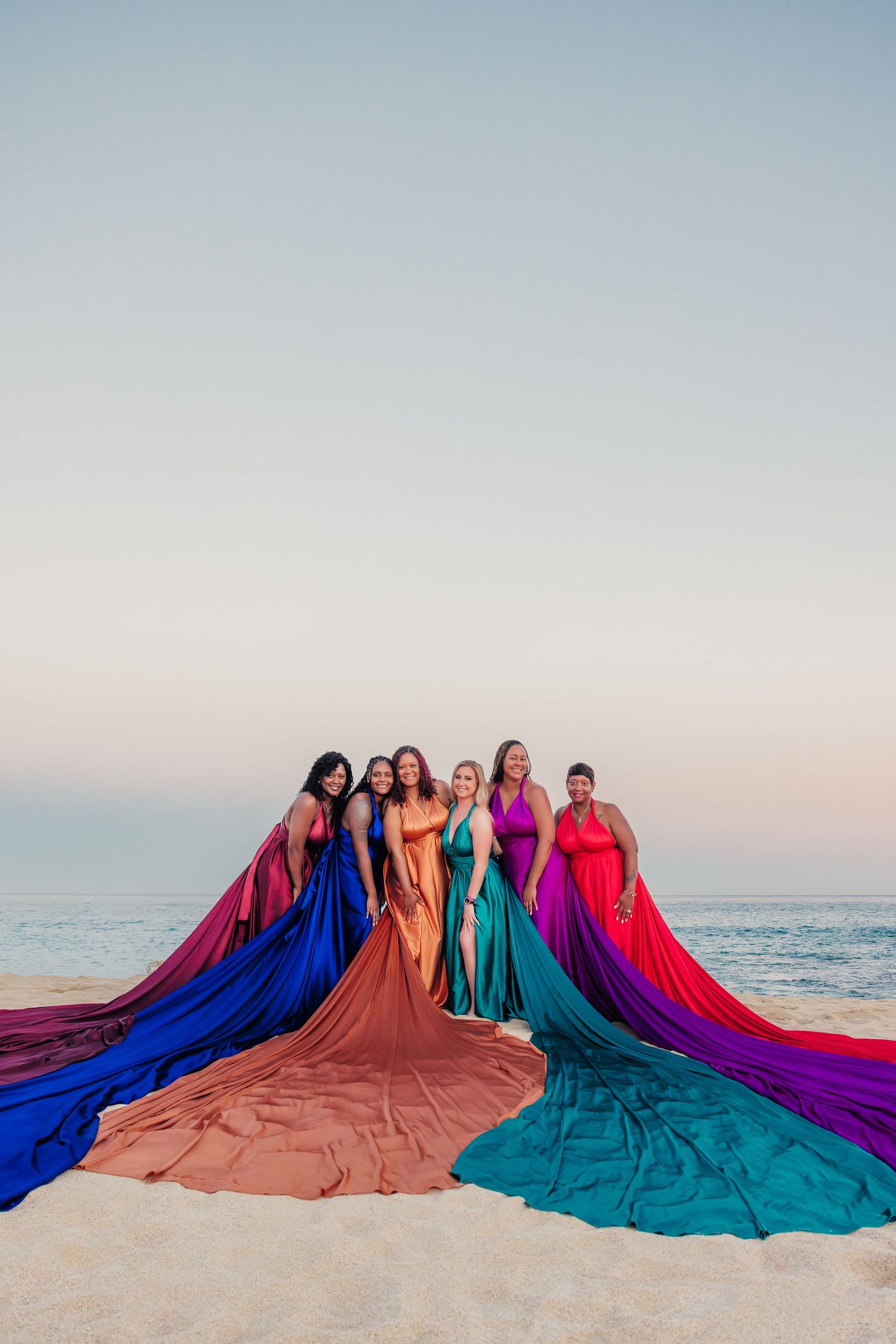 Six women in colorful long flowing gowns pose together on beach at dusk.