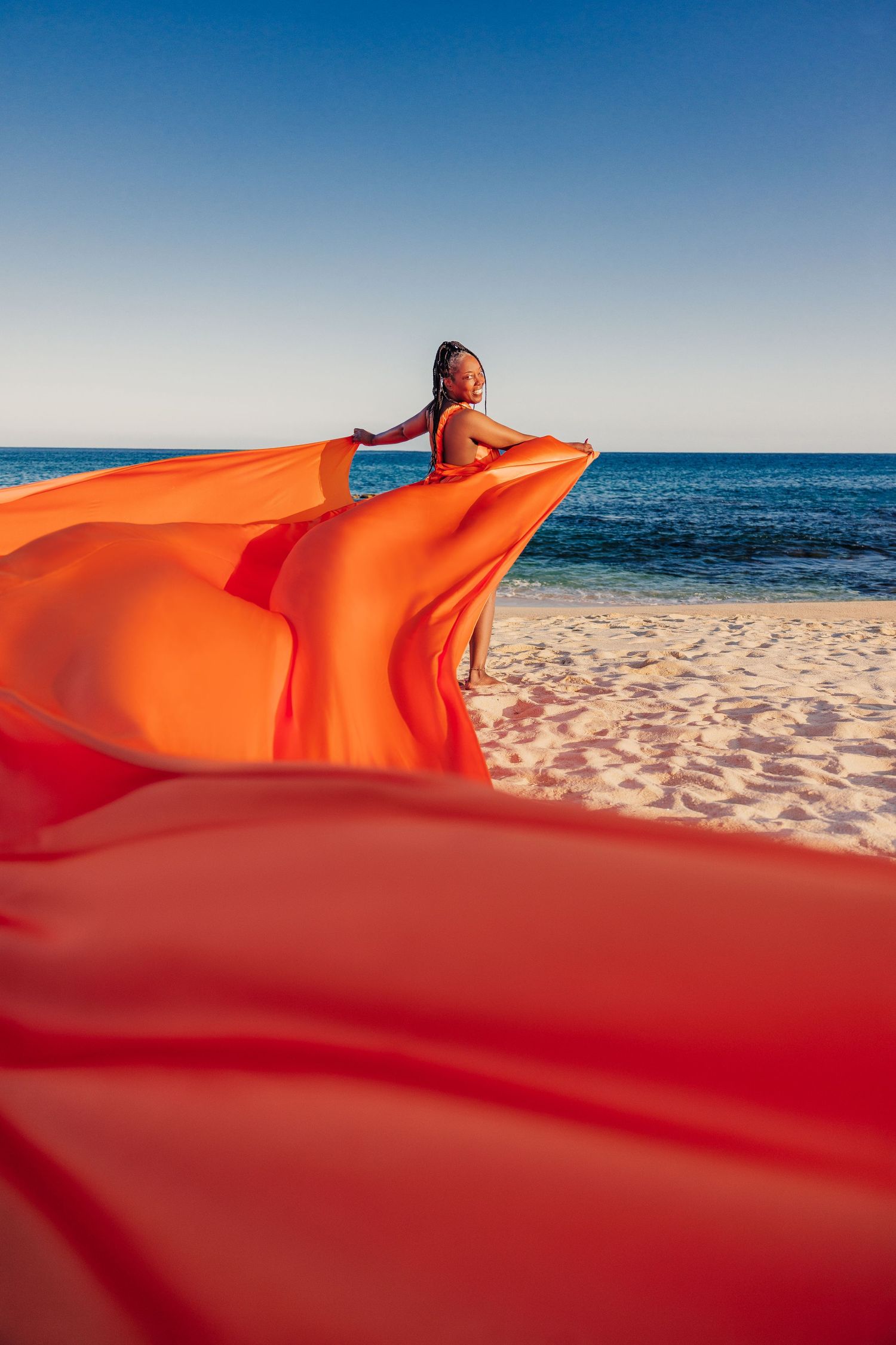 Woman in vibrant orange flying dress poses on sandy beach with blue ocean backdrop.