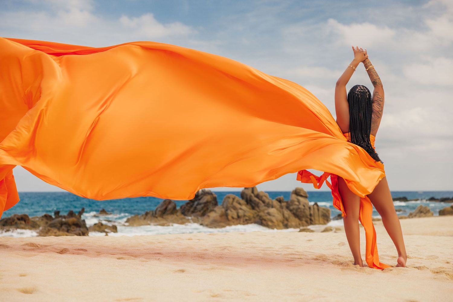 Woman on sandy beach holding flowing orange fabric billowing in the wind with rocky coastline in background.