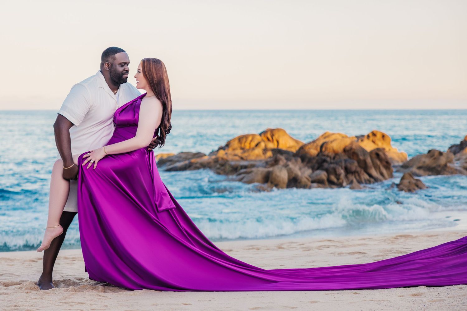 Couple in romantic beach pose, woman in flowing purple gown, man in white shirt, ocean rocks backdrop at sunset.