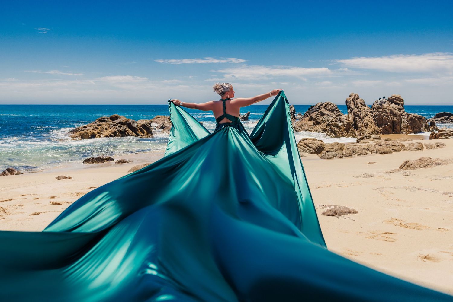 Woman in teal flowing gown poses on sandy beach with rocky coastline and blue ocean waves in background.