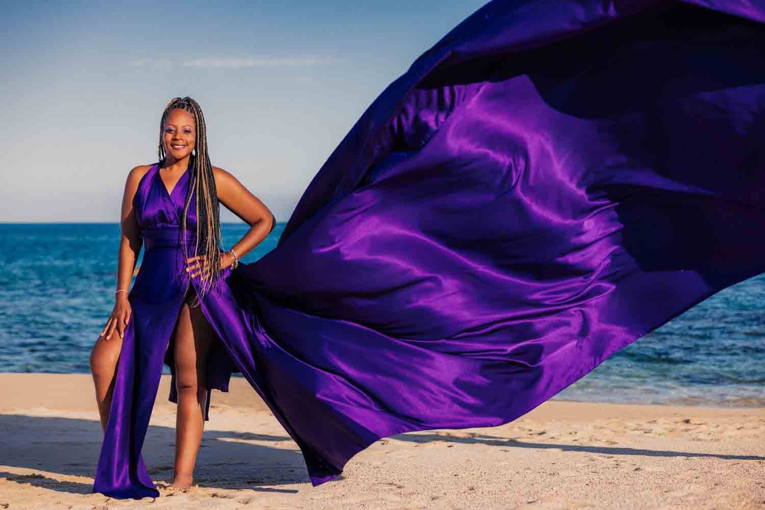 Woman in flowing purple satin gown poses on sandy beach with fabric billowing dramatically in ocean breeze.
