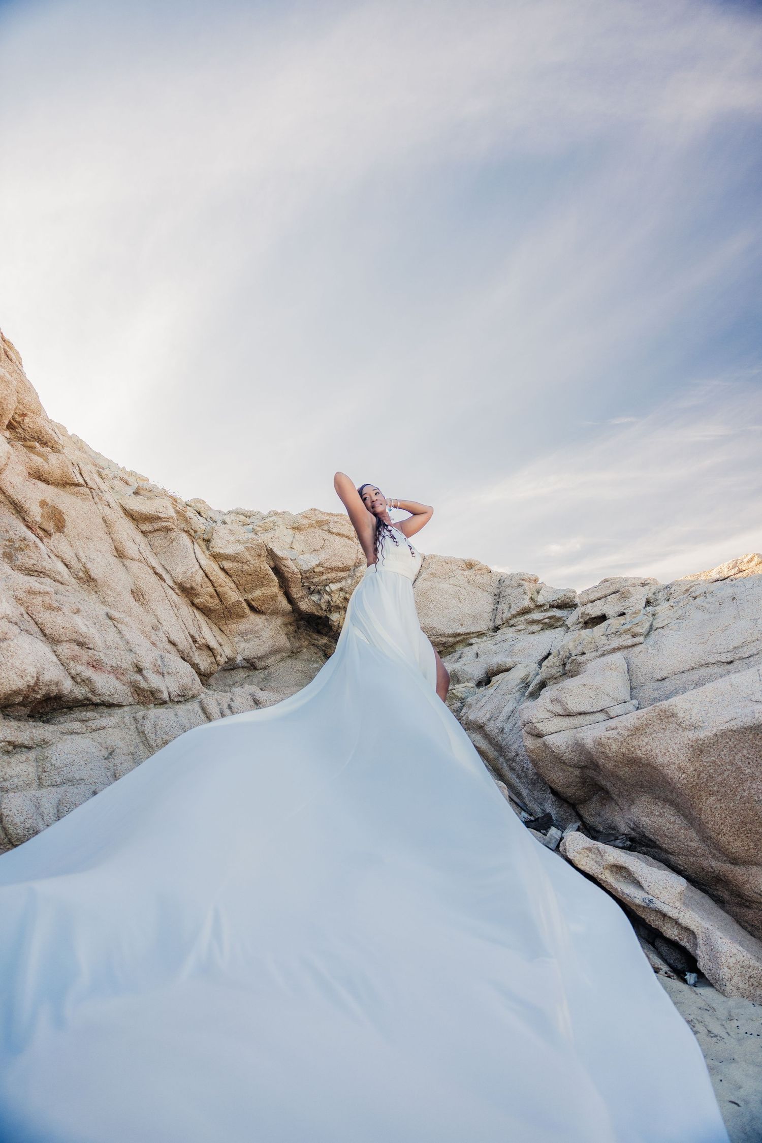 Bride in flowing white gown poses among rocky coastal cliffs with dramatic billowing train against a blue sky.
