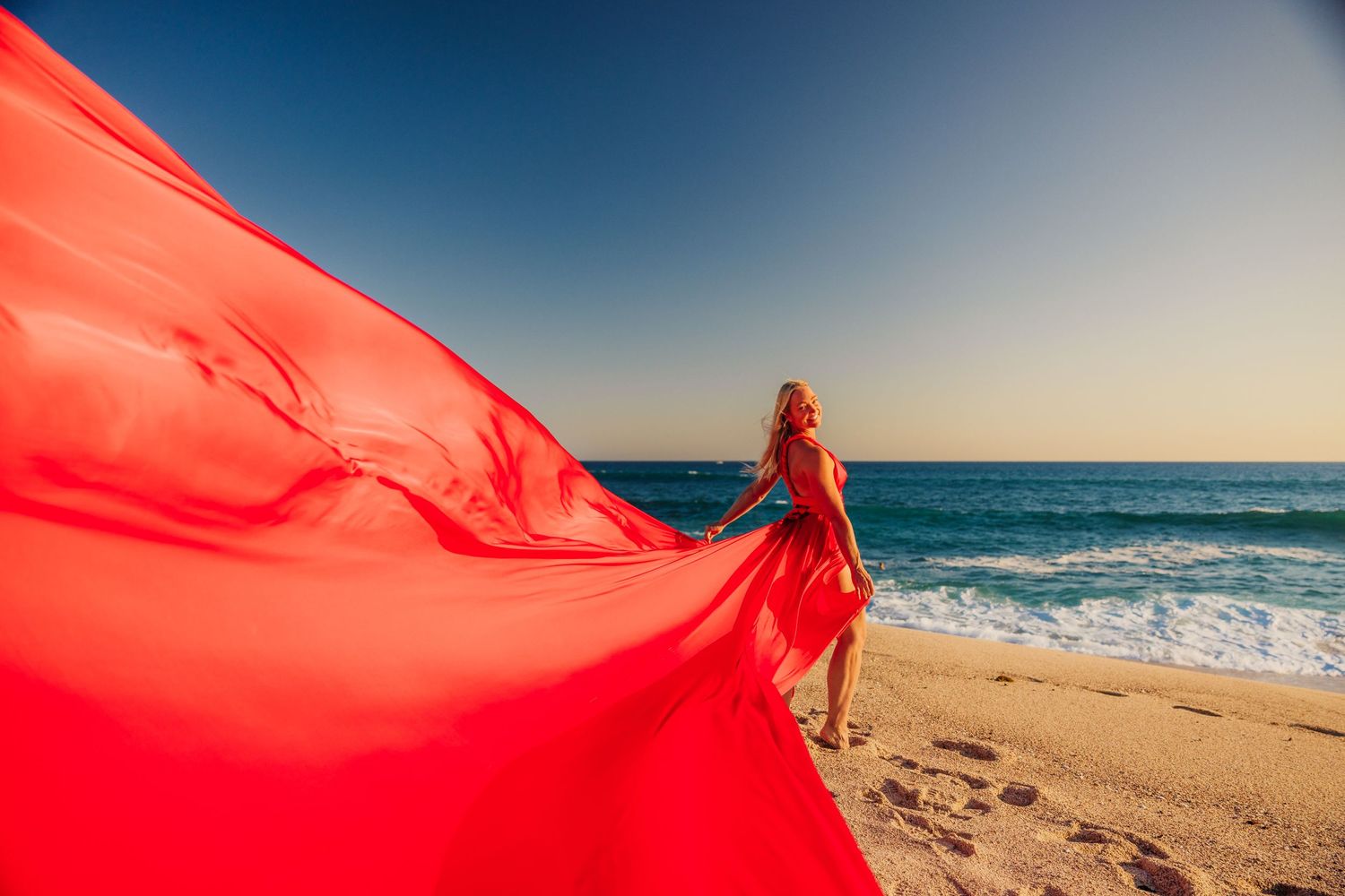 Woman in flowing red dress on sandy beach with ocean waves and clear blue sky backdrop.