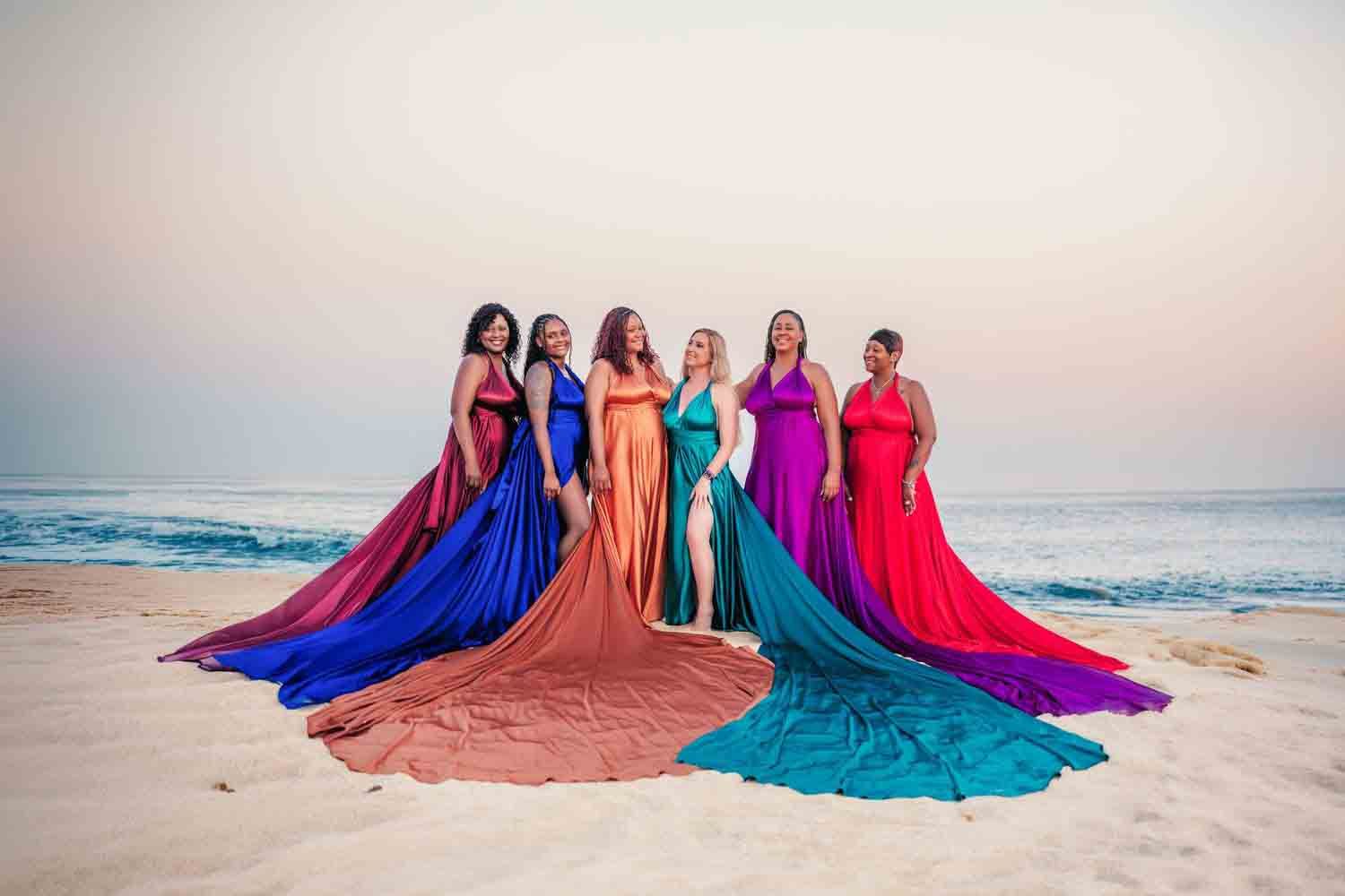 Seven women in vibrant colorful gowns pose gracefully on a beach at sunset with ocean waves in the background.