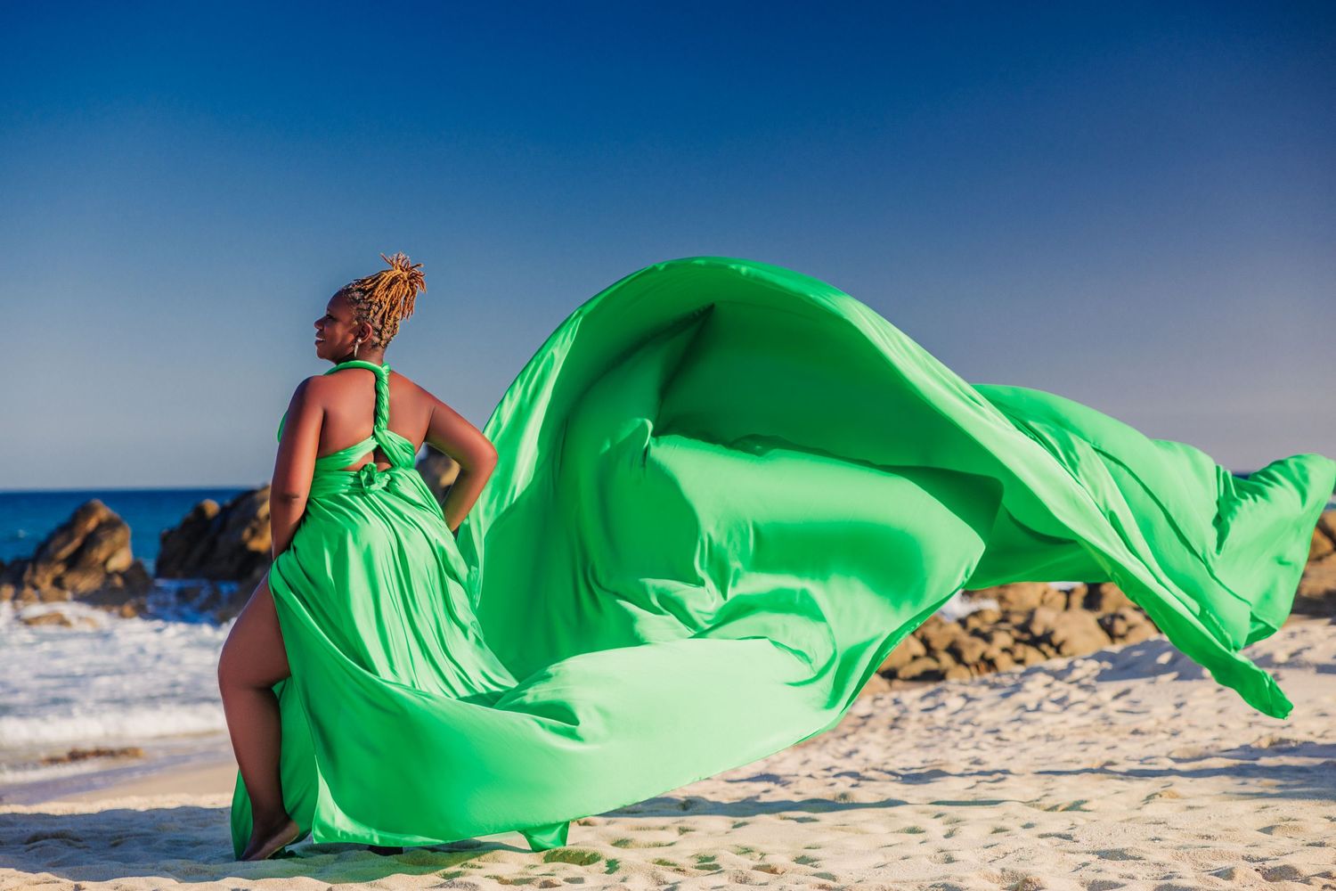 Woman in flowing green gown on sandy beach, fabric billowing in wind against blue sky and rocky shore.