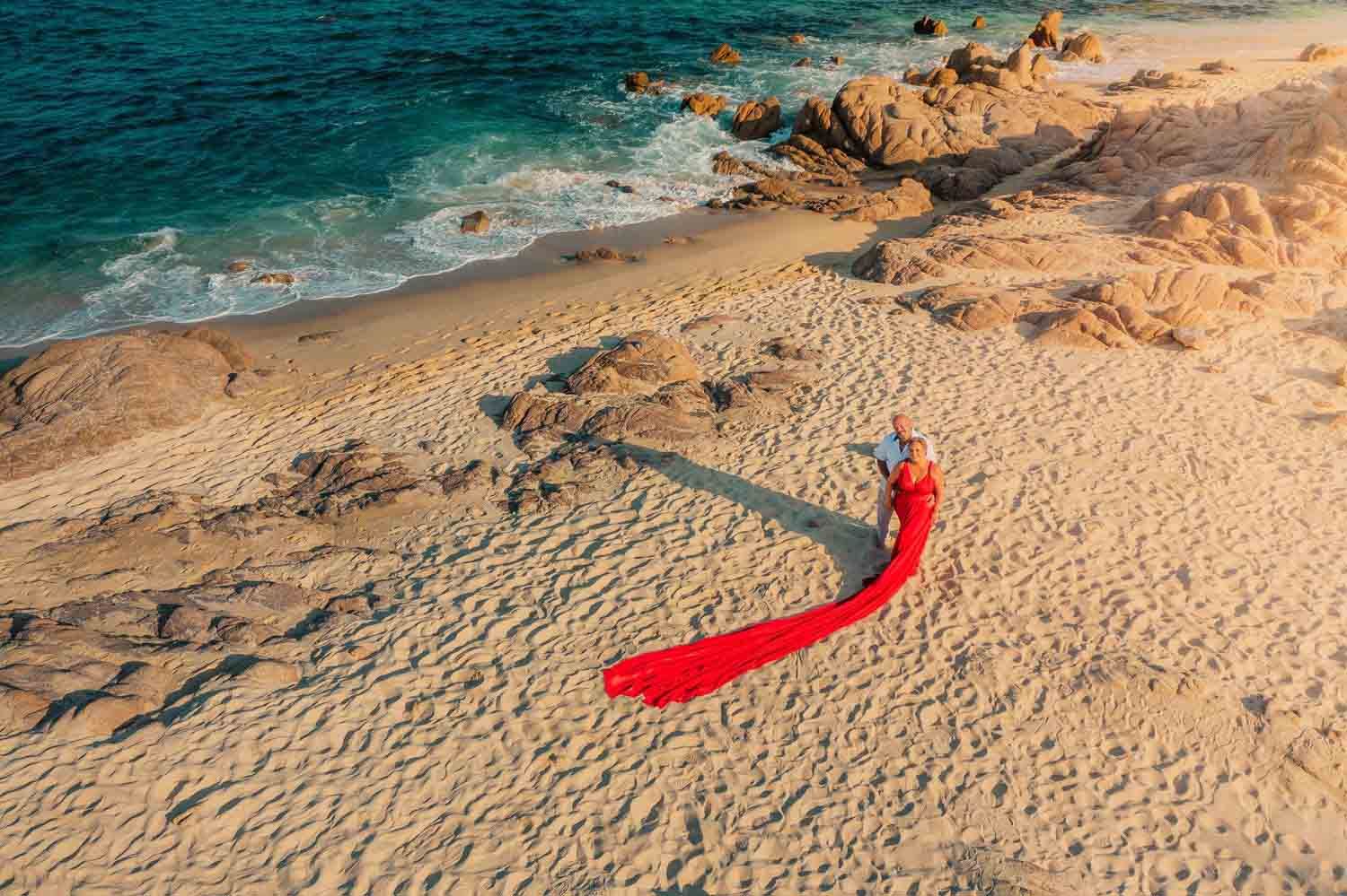 Aerial view of a woman in a flowing red dress on a sandy beach with turquoise waters and rocky shores.