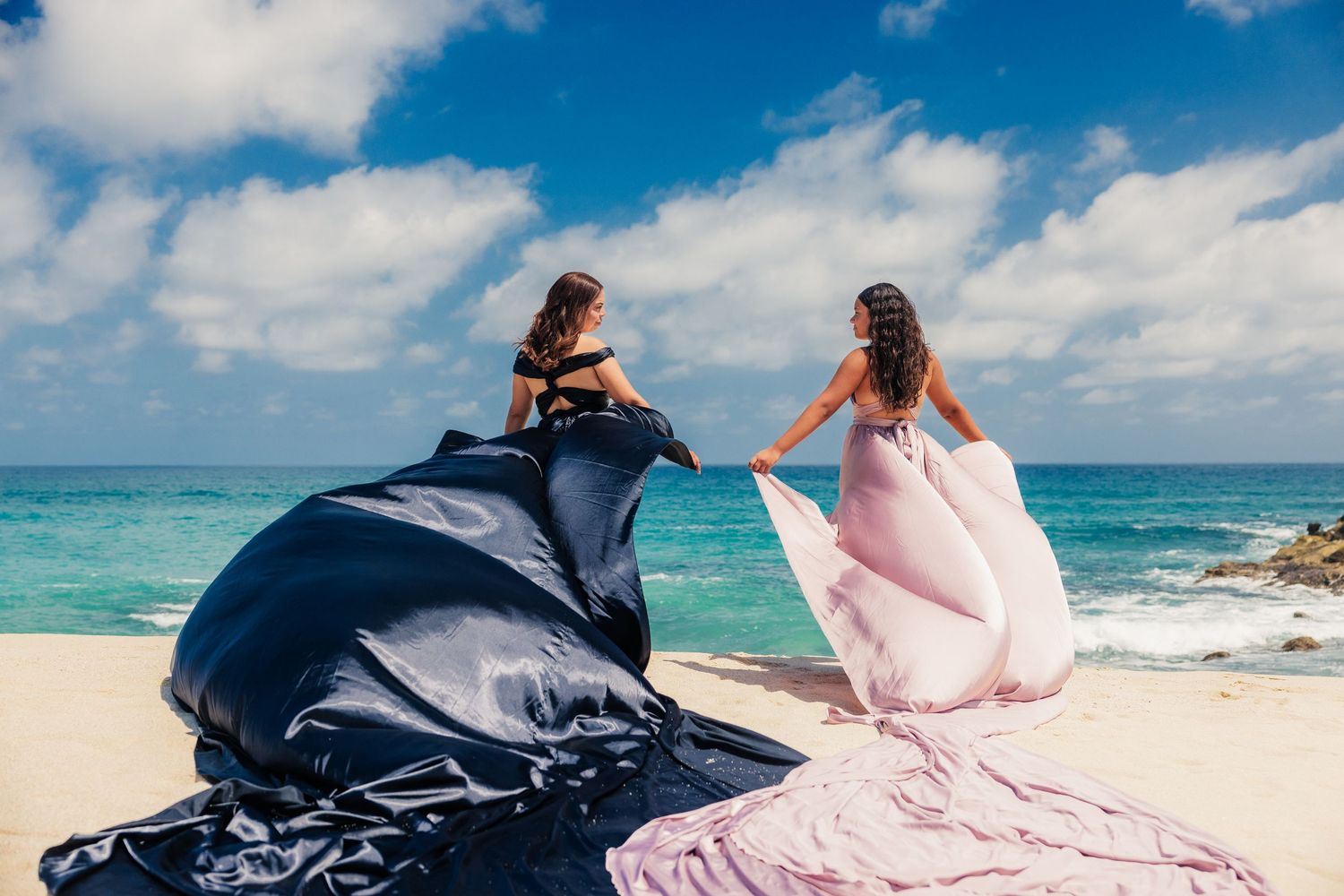 Two women in flowing gowns on a tropical beach, dresses billowing in the wind against a blue sky.