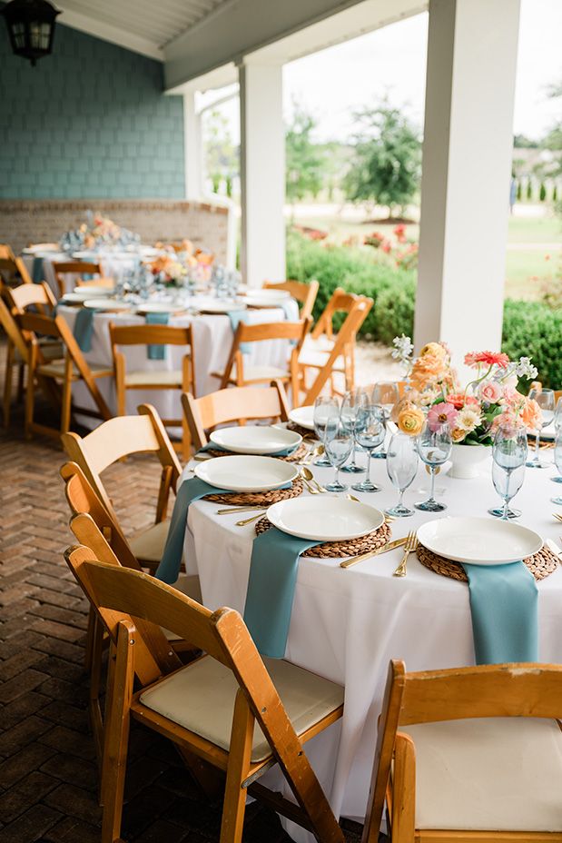 Elegant wedding reception tables set with white linens, blue runners, and wooden chairs in a covered outdoor venue.
