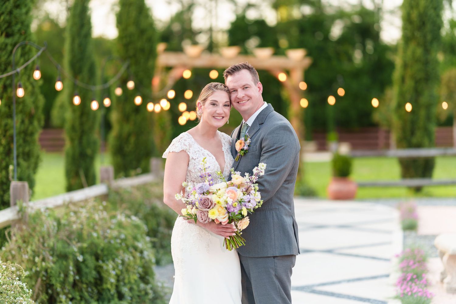 A couple embraces at an elegant outdoor wedding with string lights and cypress trees in the background.
