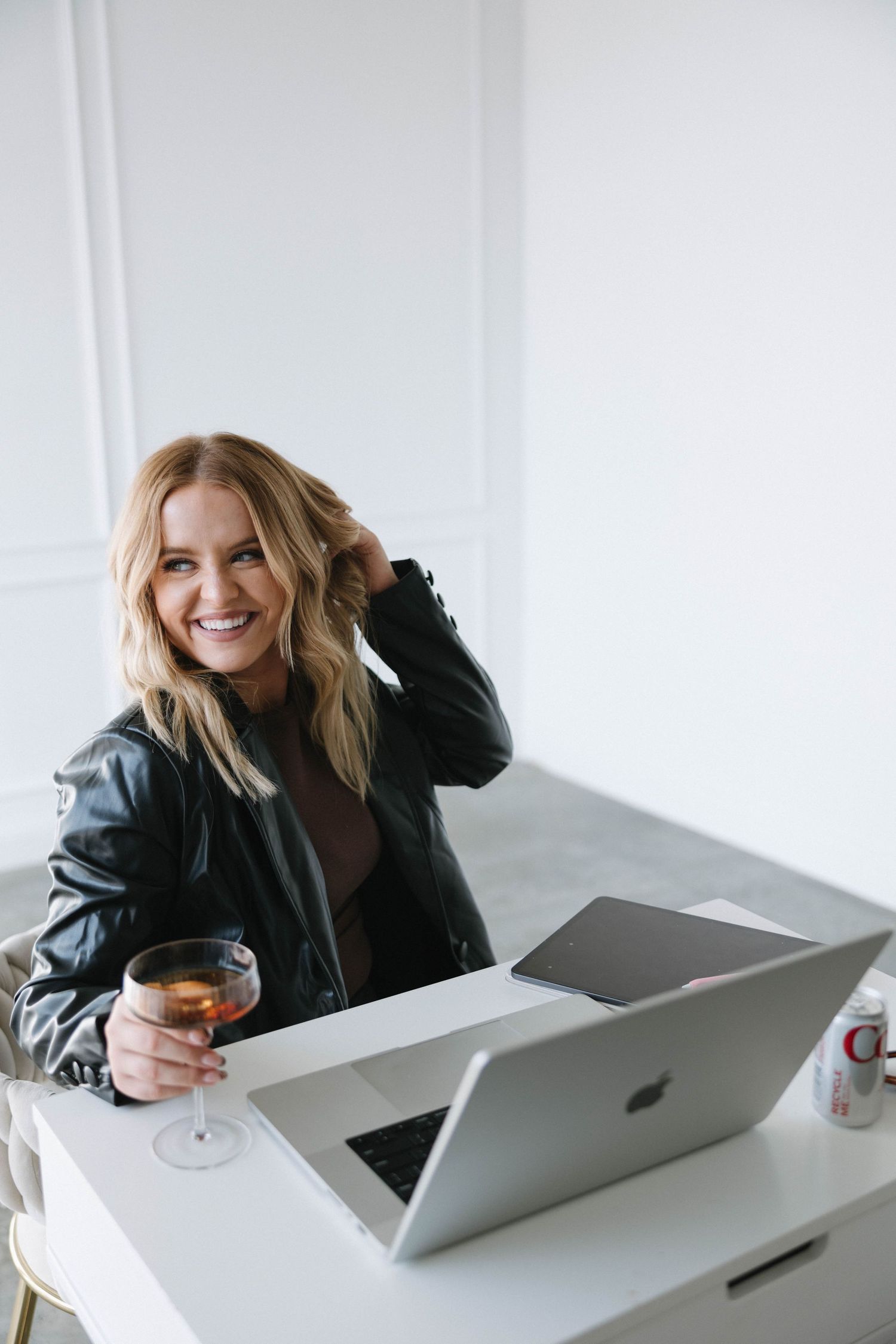 Person working on MacBook laptop at white desk with beverage in casual leather jacket.