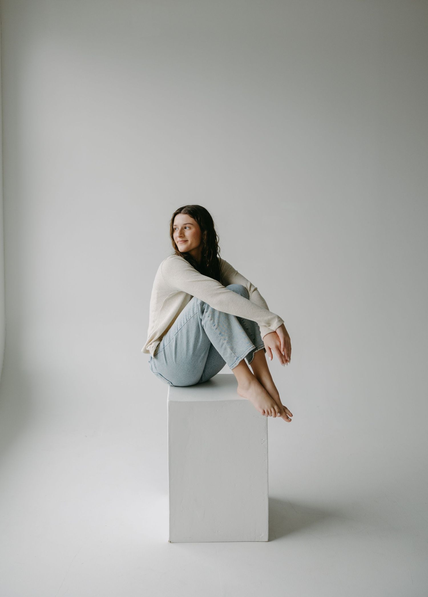 Minimalist portrait of a person in casual wear sitting on a white cube against grey background.