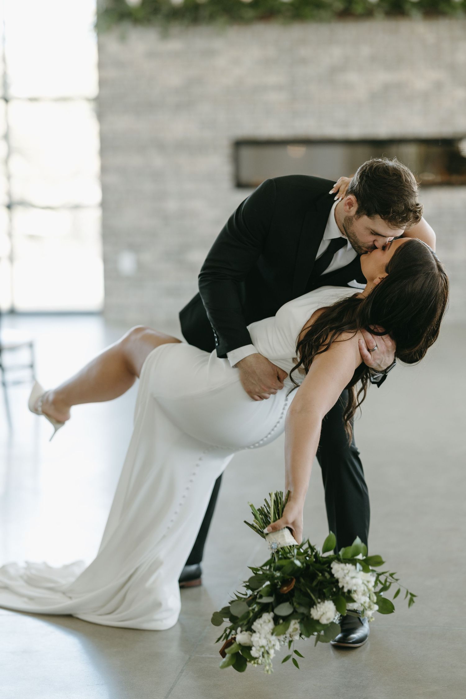 Romantic wedding dip pose with bride holding white floral bouquet in modern venue.