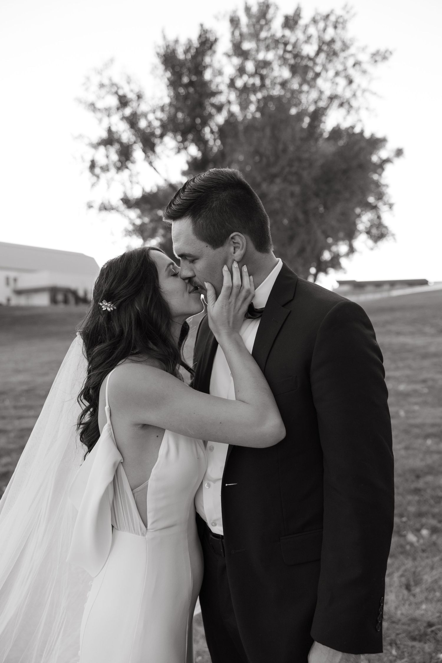 A black and white outdoor portrait captures a romantic kiss between newlyweds.
