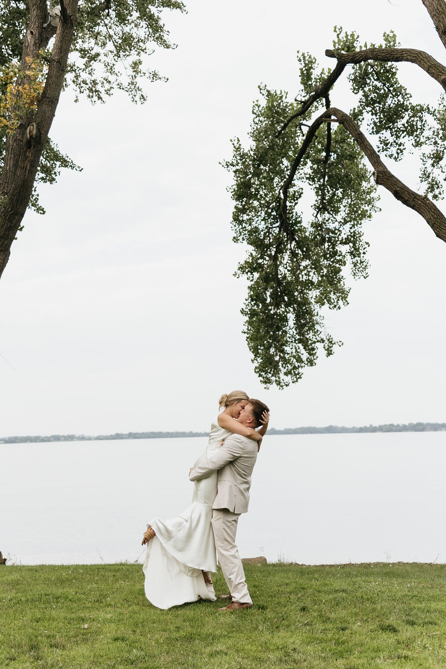 A couple embraces on a grassy lawn beside a lake during their wedding celebration.
