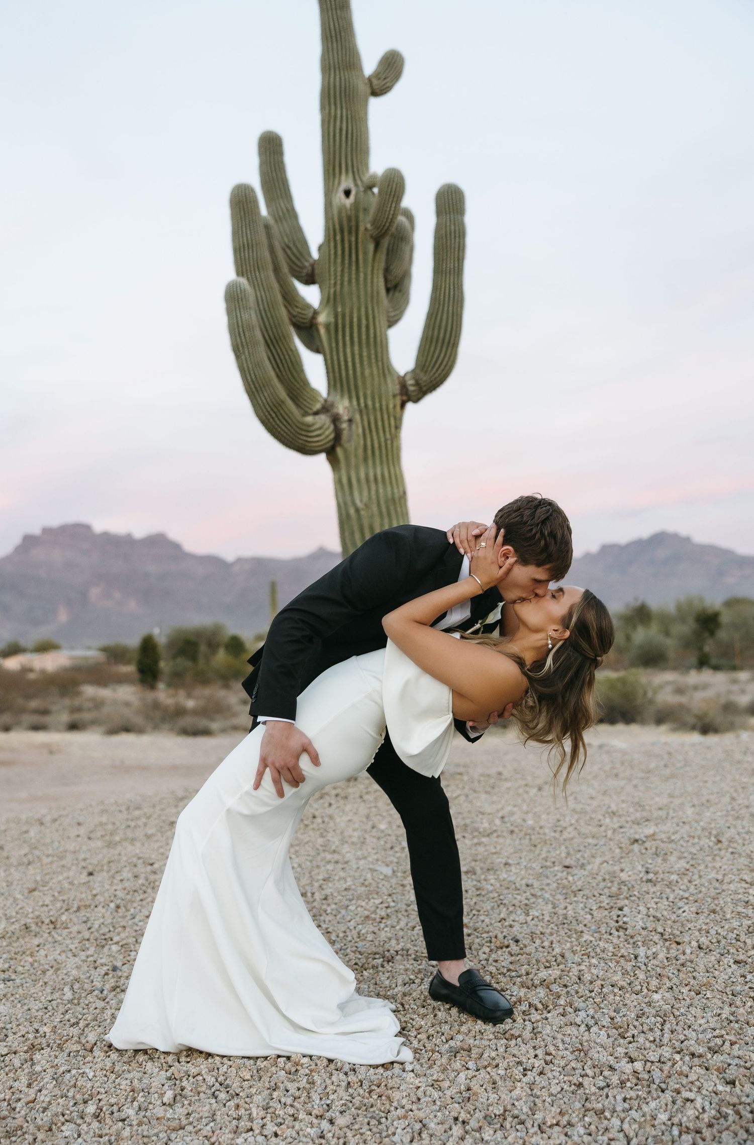 A dramatic wedding kiss pose in front of a towering saguaro cactus in the desert at dusk.