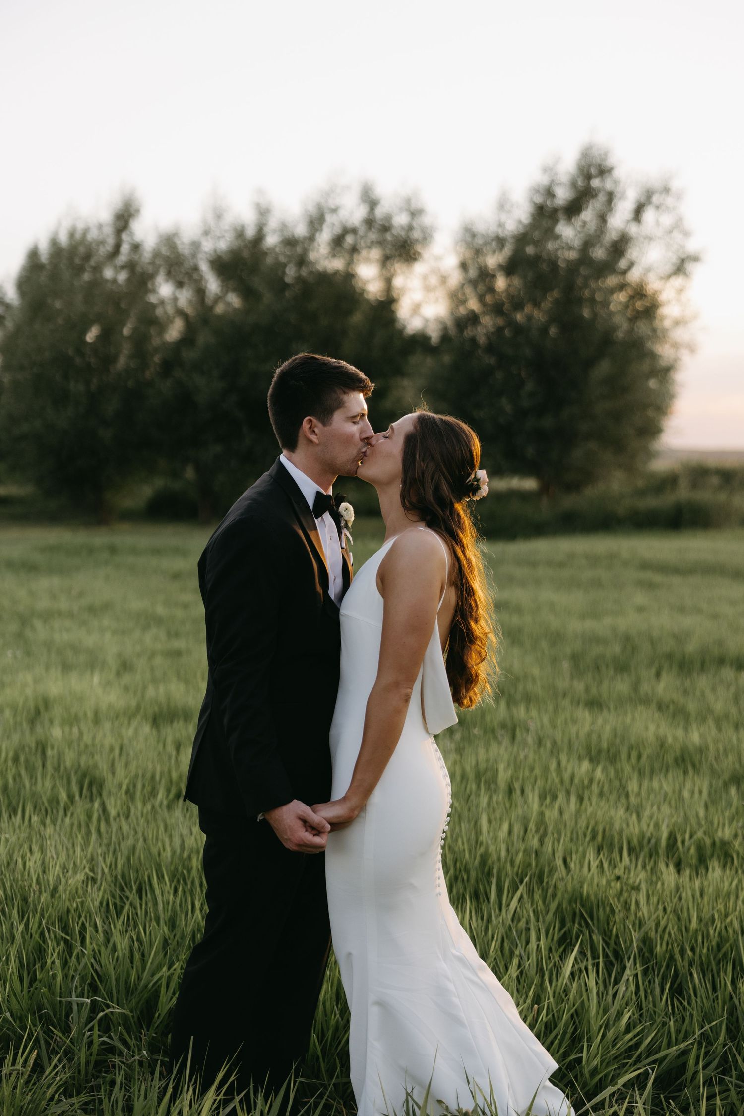 Romantic wedding photo of couple kissing in flowing grass field at sunset.