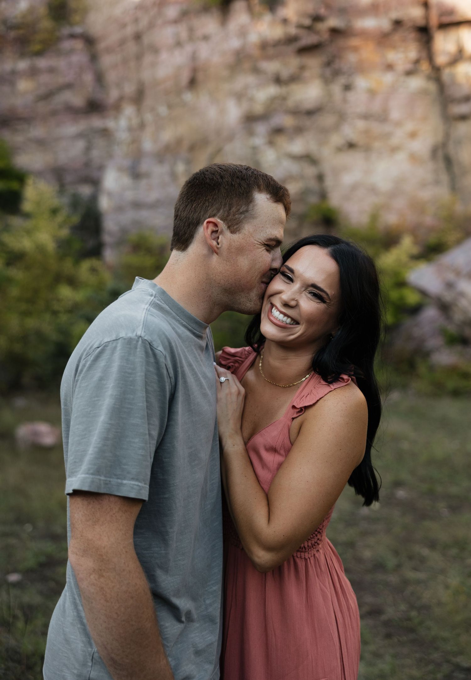 Candid romantic moment captured between couple laughing against rocky cliff background.