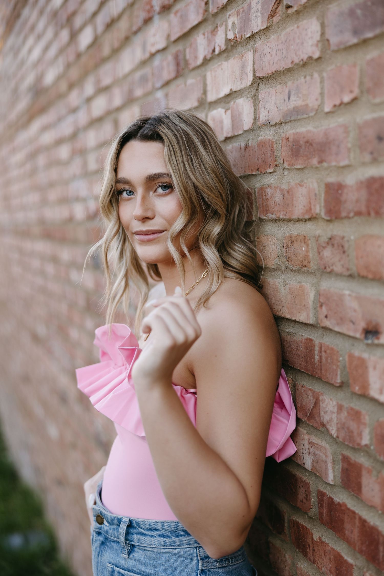 Young woman in pink top and jeans poses against brick wall.