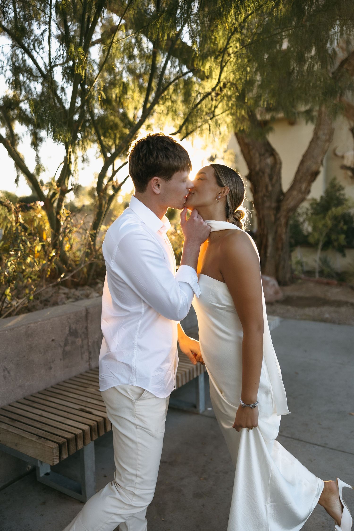 Romantic sunset kiss between couple in all-white outfits under a willow tree.