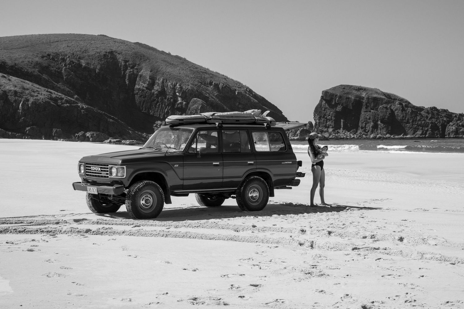 Black and white photo of a 4WD vehicle parked on a sandy beach with a person standing beside it and mountains behind.