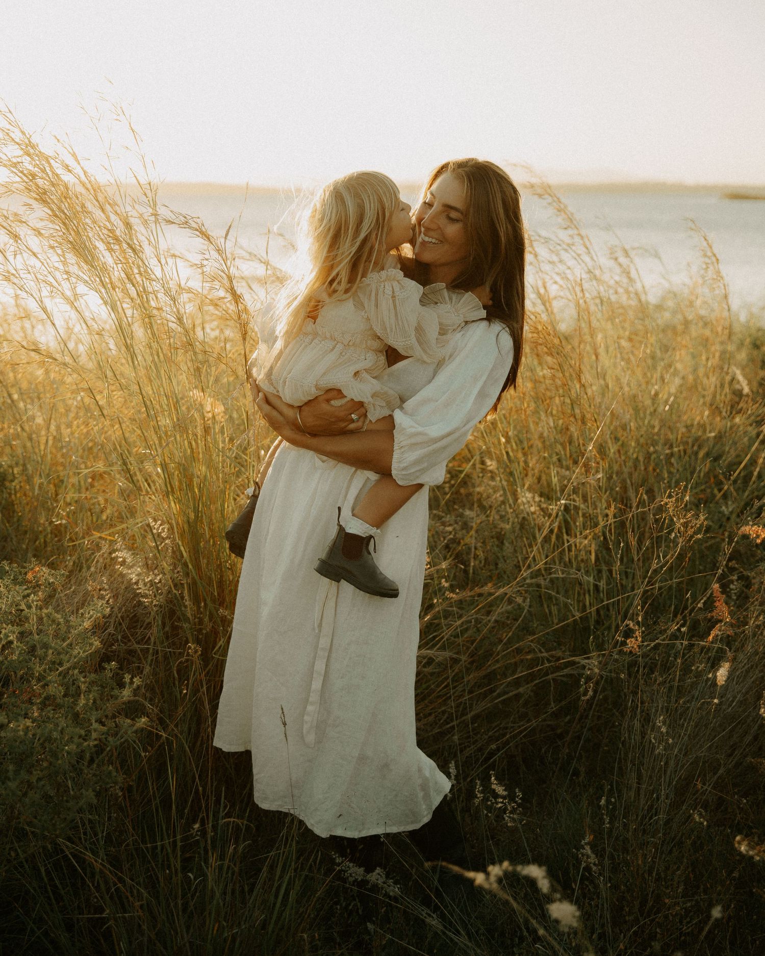 A mother in a white dress holds her young child in golden tall grass near water at sunset.